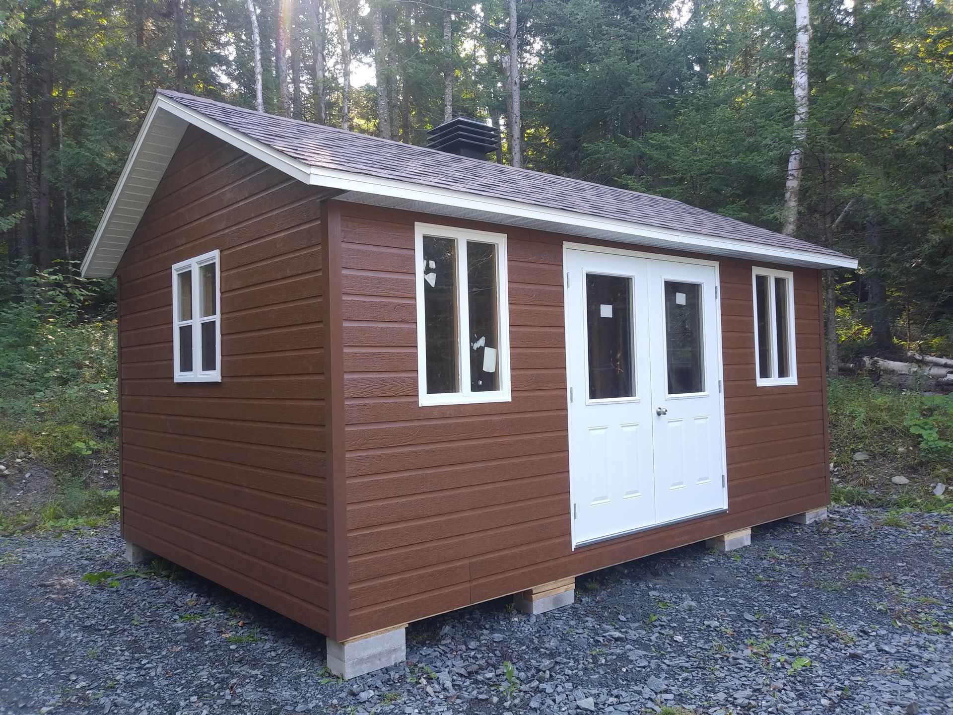 Abri de jardin marron avec des boiseries, des portes et des fenêtres blanches, sur des blocs de béton, situé dans une zone boisée.