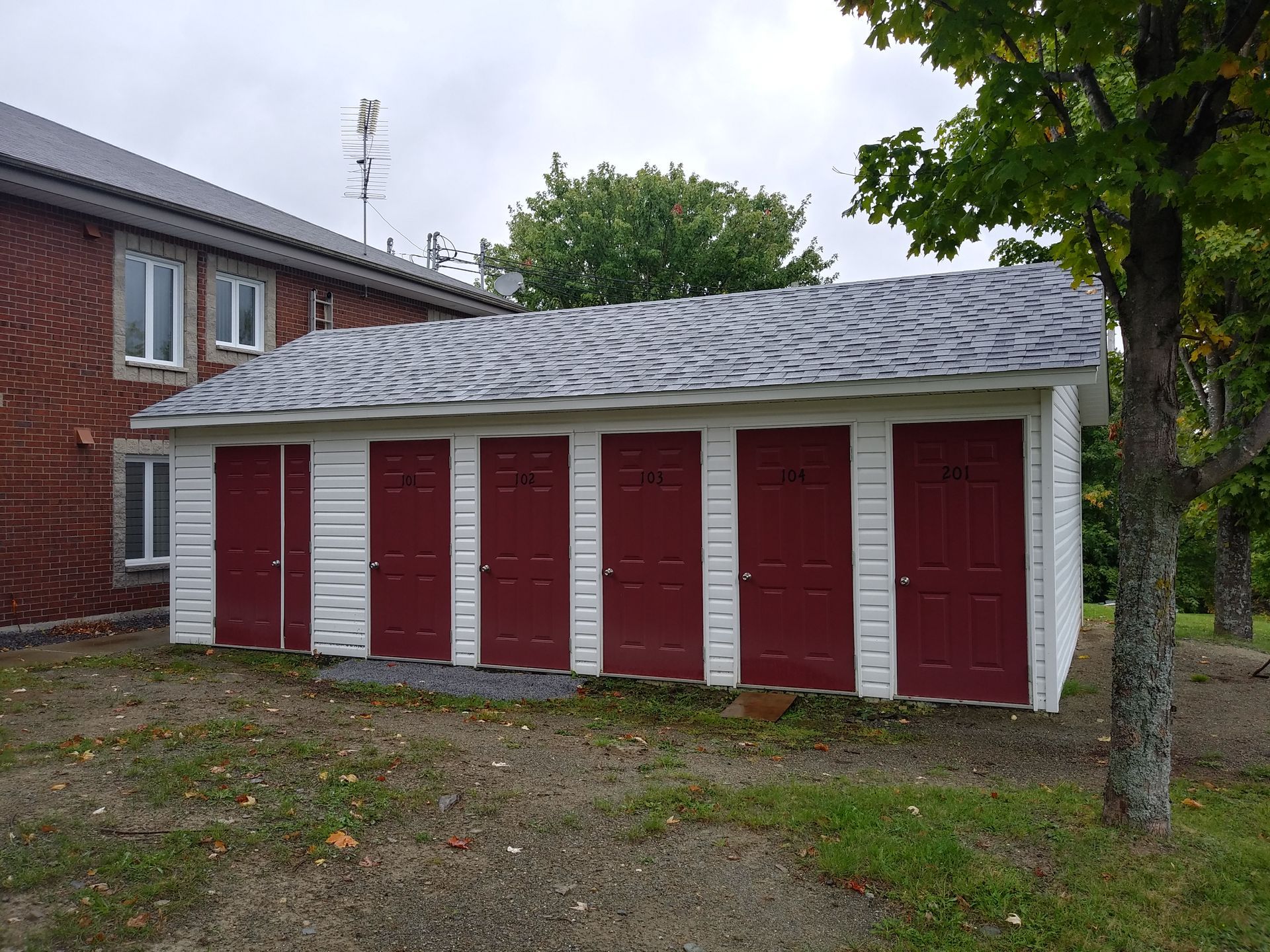 Garage rouge et blanc à six portes avec toit en bardeaux gris, jouxtant un bâtiment en briques.