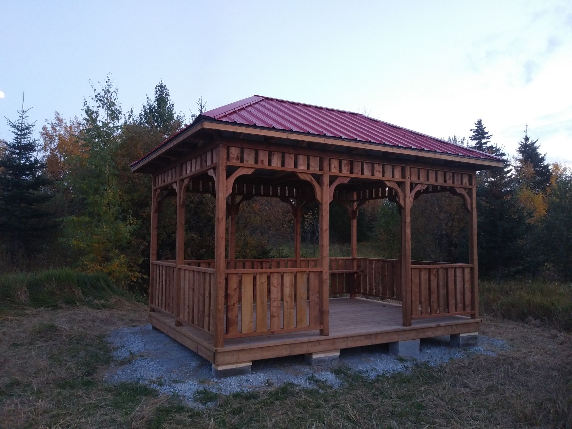 Un kiosque en bois au toit rouge se dresse dans un champ herbeux, entouré d'arbres.