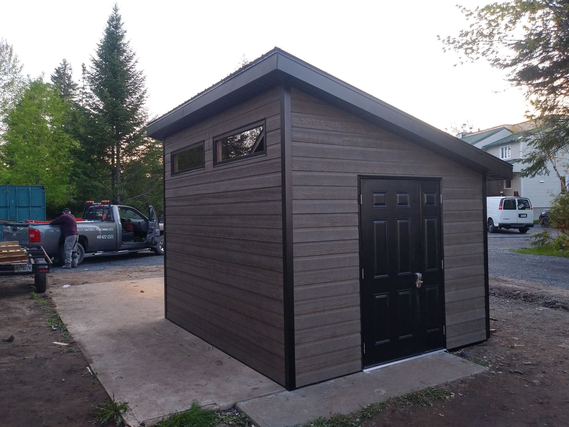 Abri marron avec porte, fenêtres et moulures noires sur une dalle de béton ; deux personnes près d'un camion en arrière-plan.