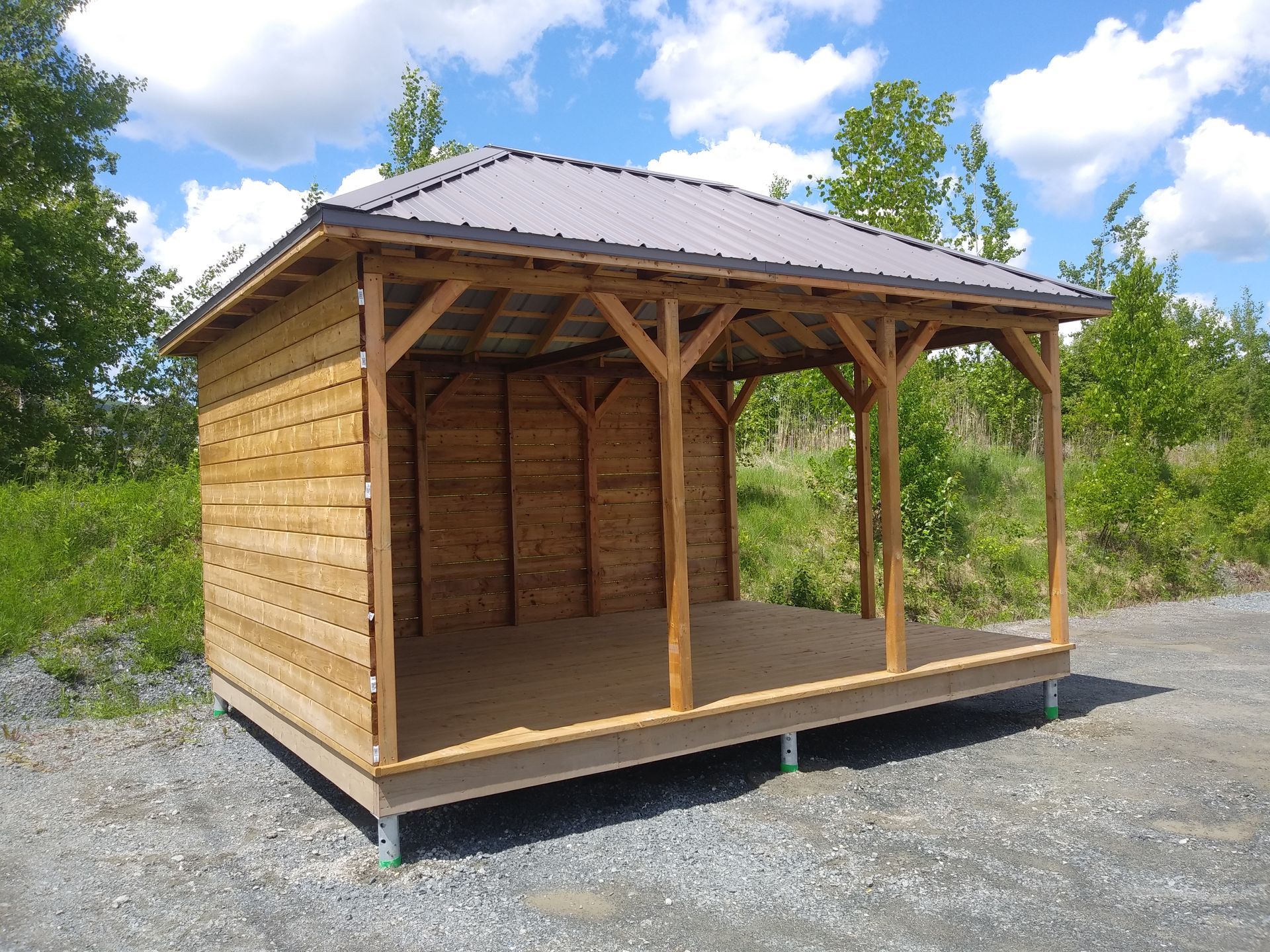 Gazebo en bois avec toit en métal, partiellement fermé. Dehors, ciel bleu.