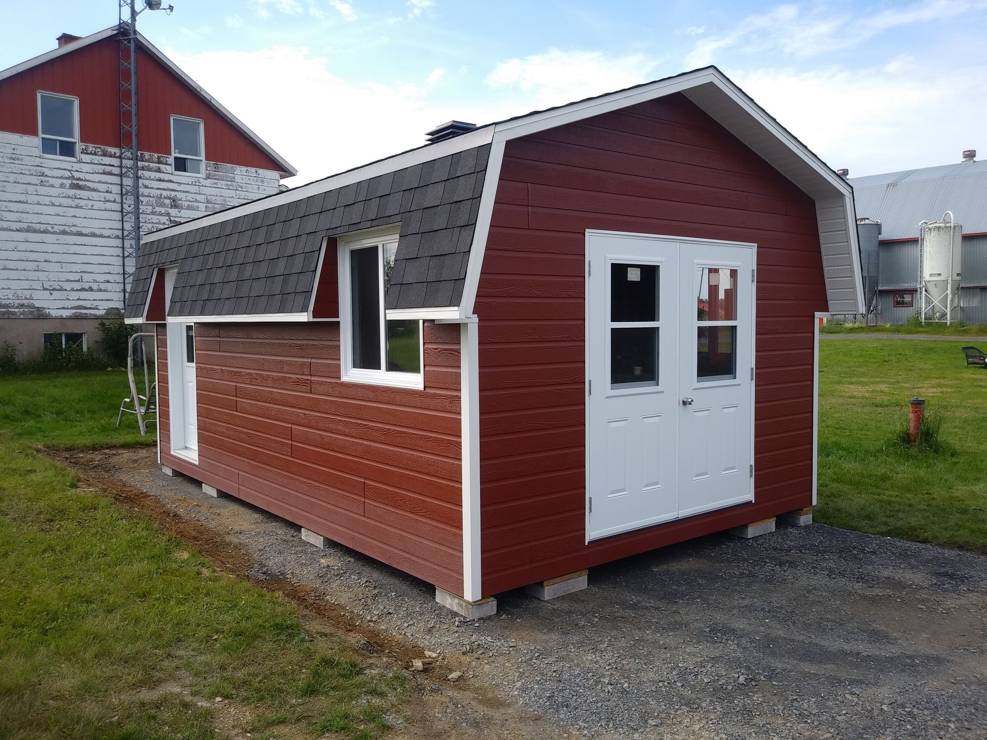 Abri de jardin rouge avec des boiseries blanches, des portes doubles et une fenêtre, posé sur du gravier.