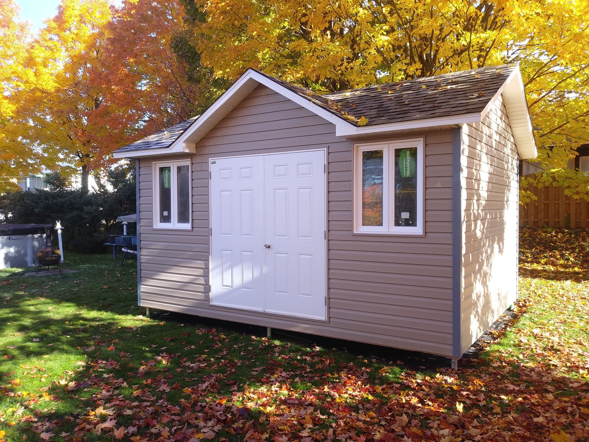 Abri de jardin beige avec portes et fenêtres blanches, situé dans une cour ornée de feuillage d'automne et d'herbe verte.