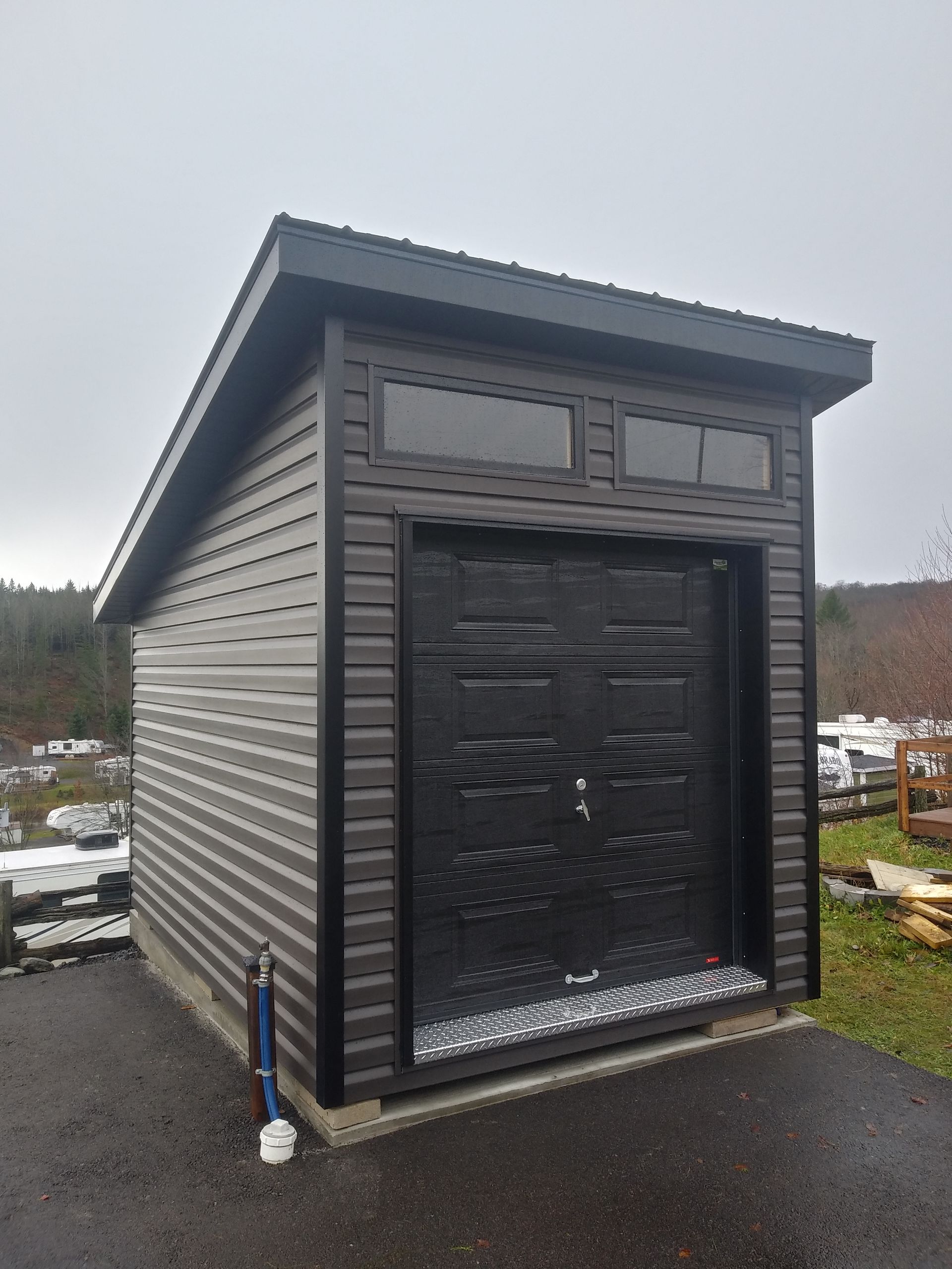 Abri de jardin brun avec porte de garage foncée, fenêtres et marche grise. Ciel couvert.