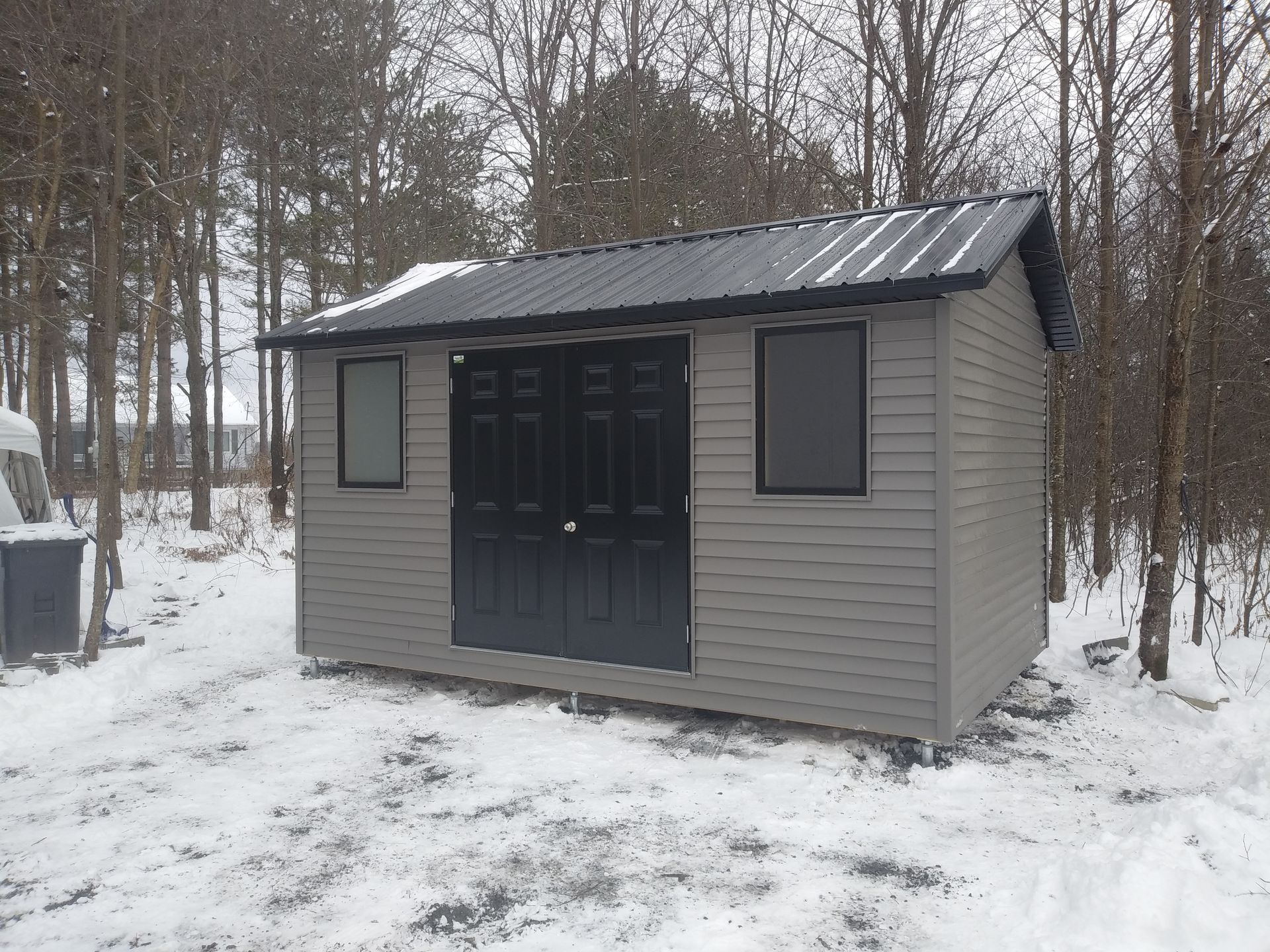 Abri gris avec portes et toit noirs dans un décor extérieur enneigé.