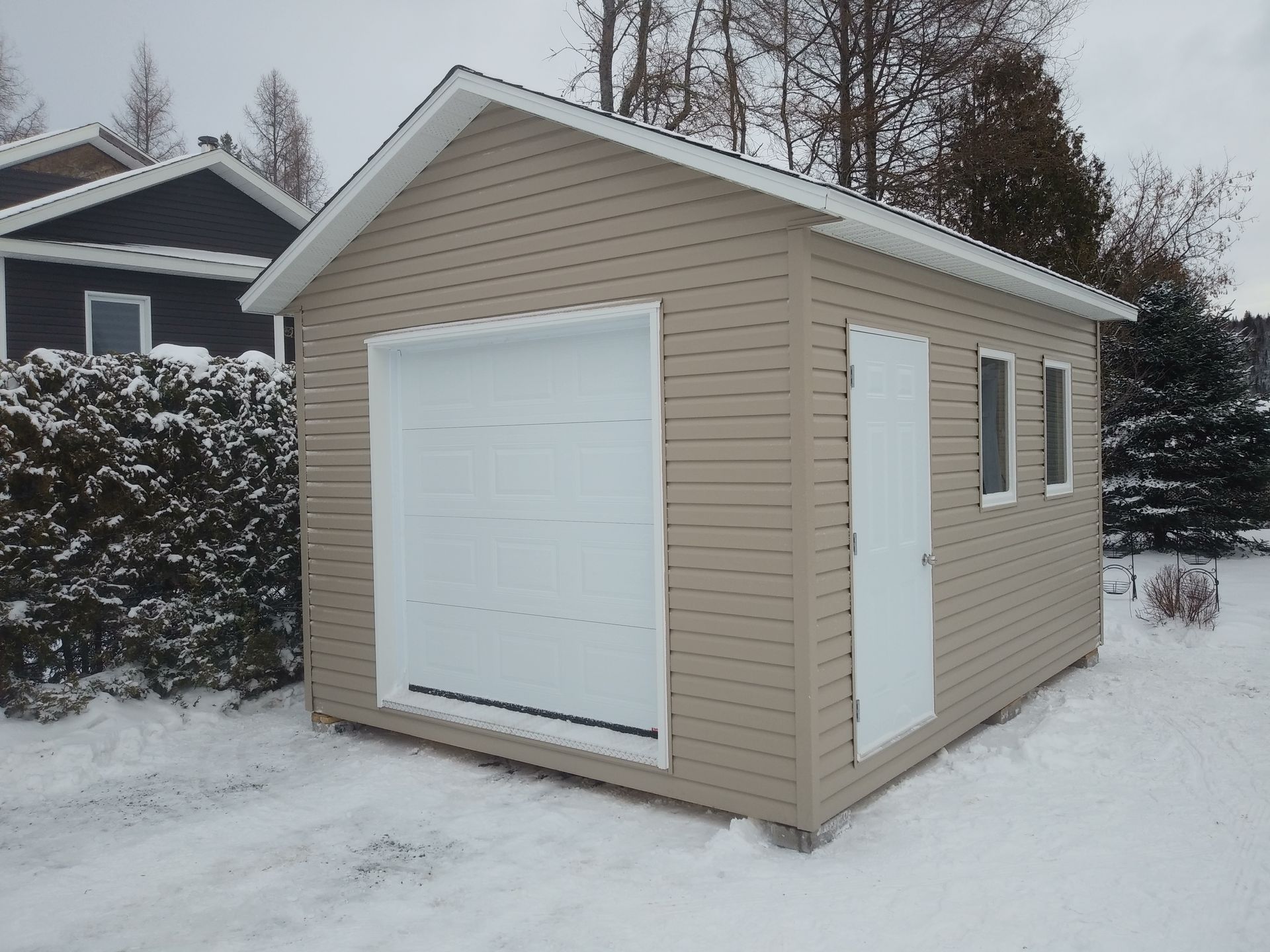 Garage beige avec encadrement de porte et de fenêtre blanc, situé dans une cour enneigée.