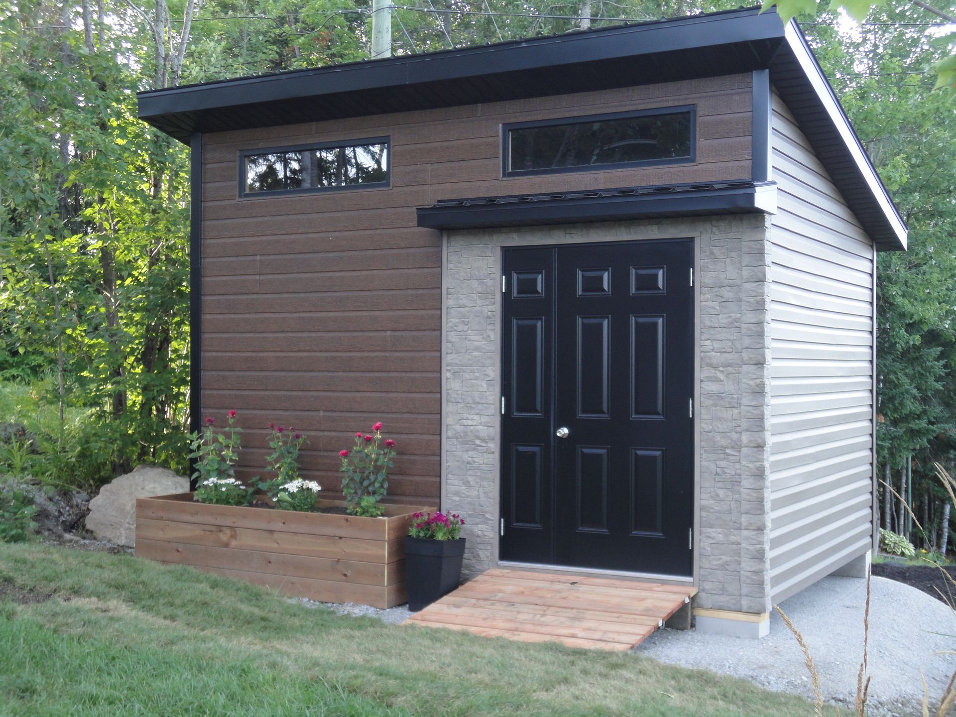 Abri de jardin moderne avec bardage marron, porte noire, petite rampe et jardinière.