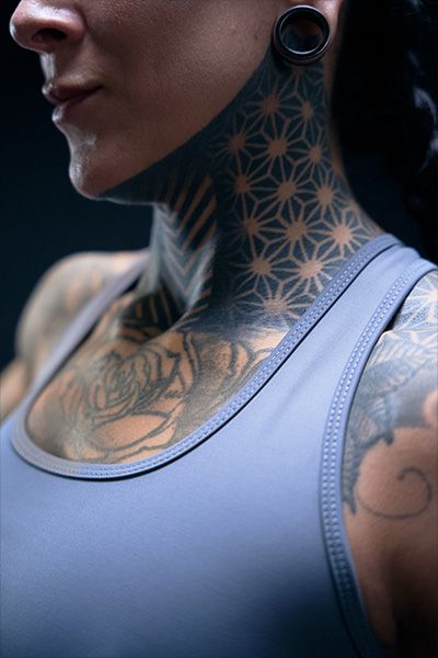 Mujer con grandes tatuajes en el cuello y el brazo, vestida con una camiseta sin mangas azul.