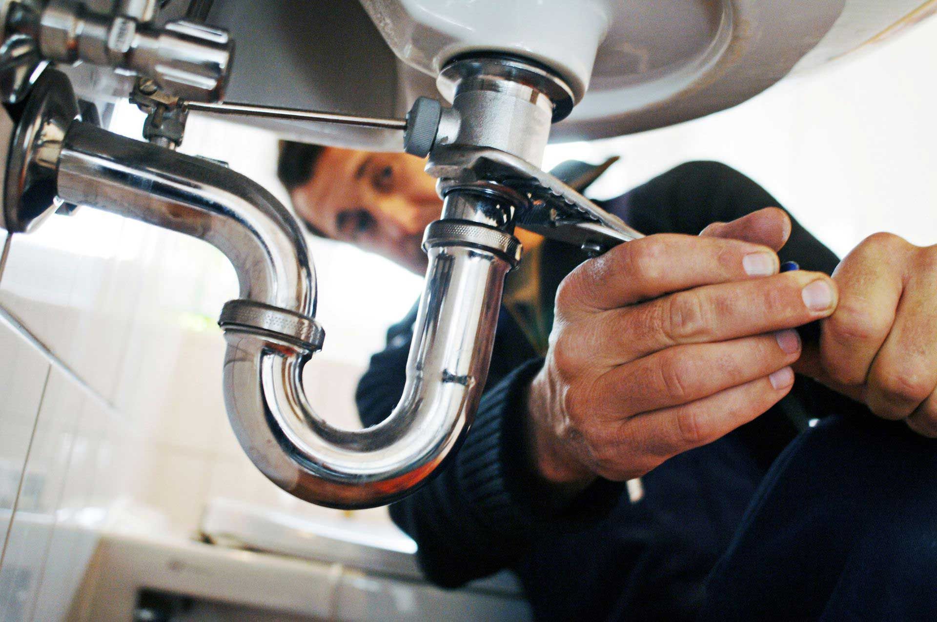 Plumber working under a sink with a wrench. Chrome pipes and white sink in a bathroom setting.