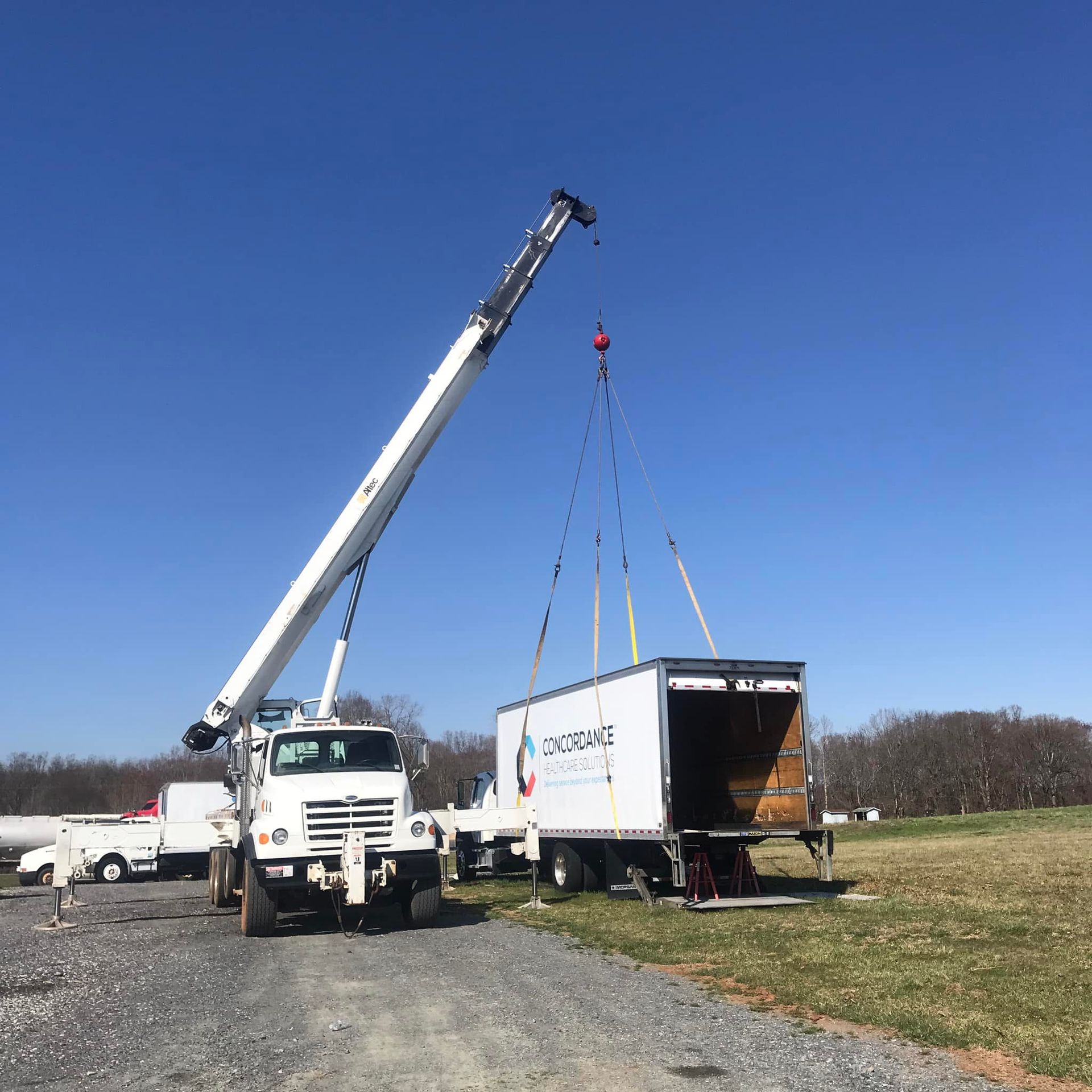 A crane lifts a white container from a flatbed truck in a field on a sunny day.