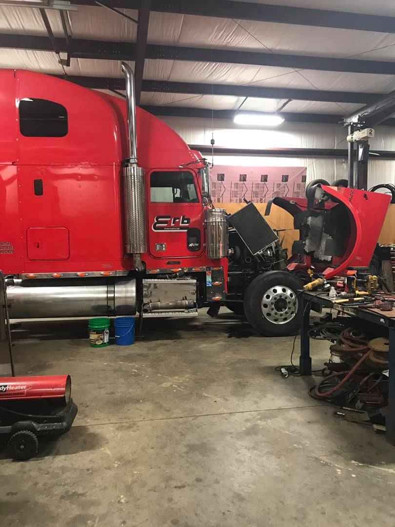 Red semi-truck in a garage with open hood, being worked on. Tools, oil, and a heater are visible.