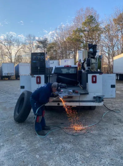 Man in blue suit welding metal on the back of a truck with a tire. Outdoors, sunny.