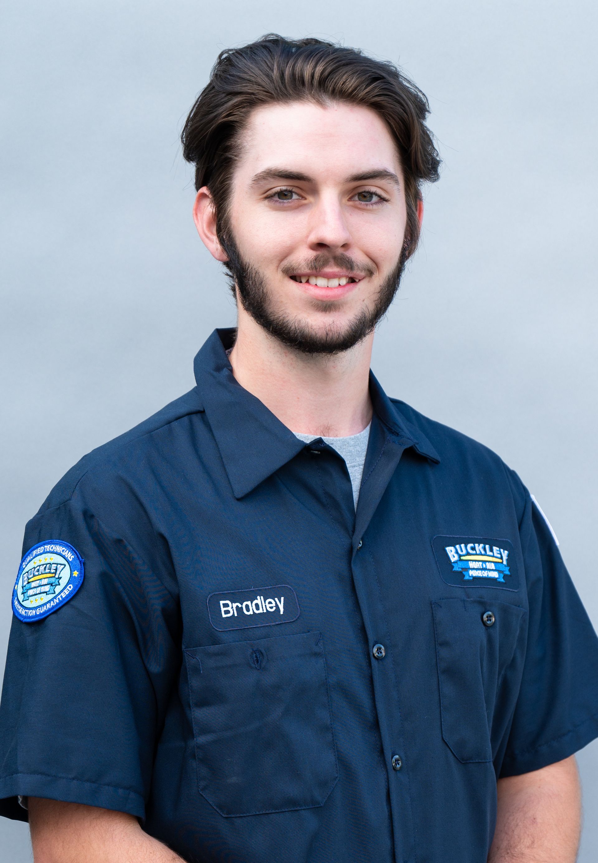 A young man with a beard is wearing a blue shirt and smiling.