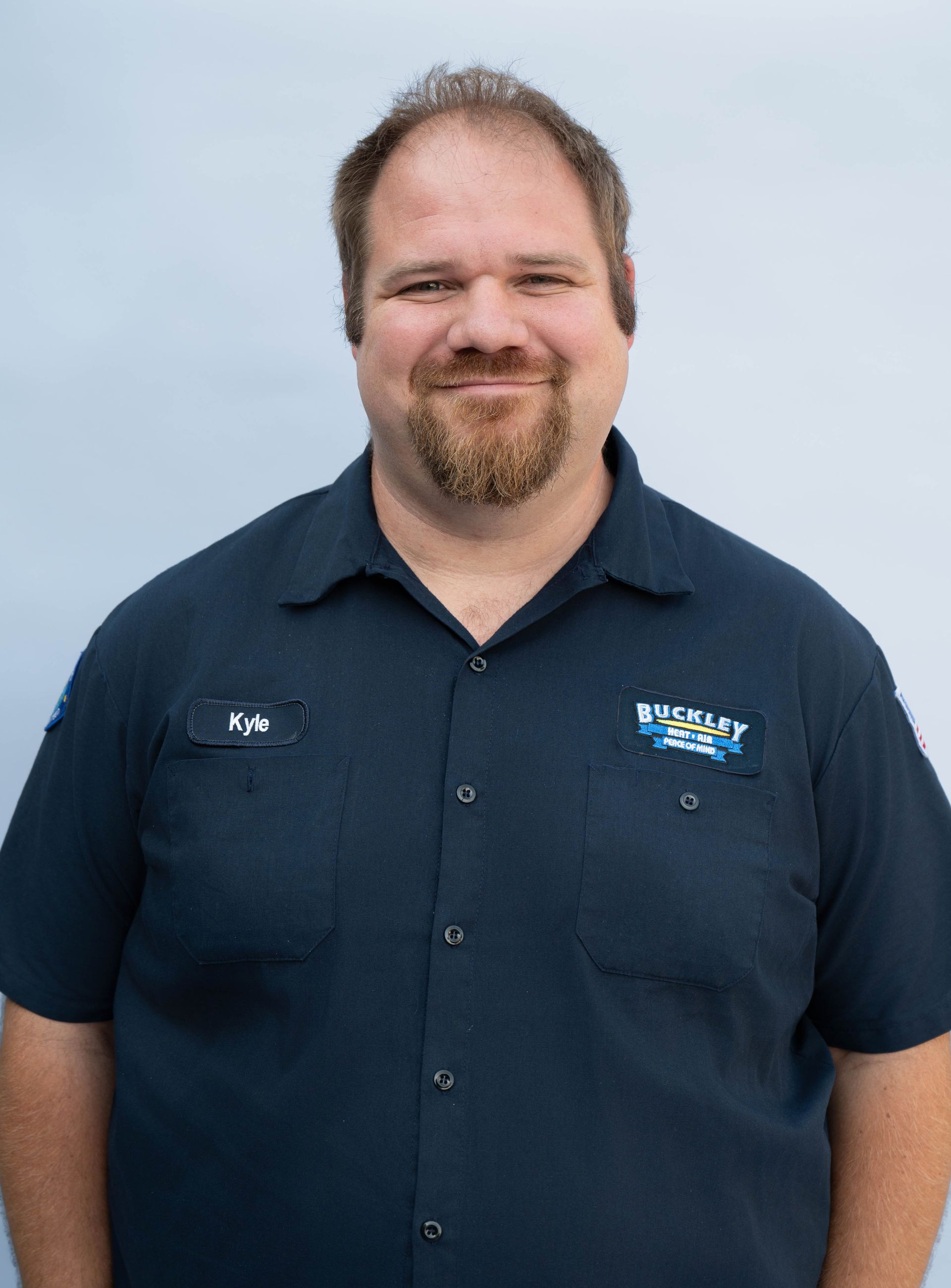 A young man with a beard is wearing a blue shirt and smiling.