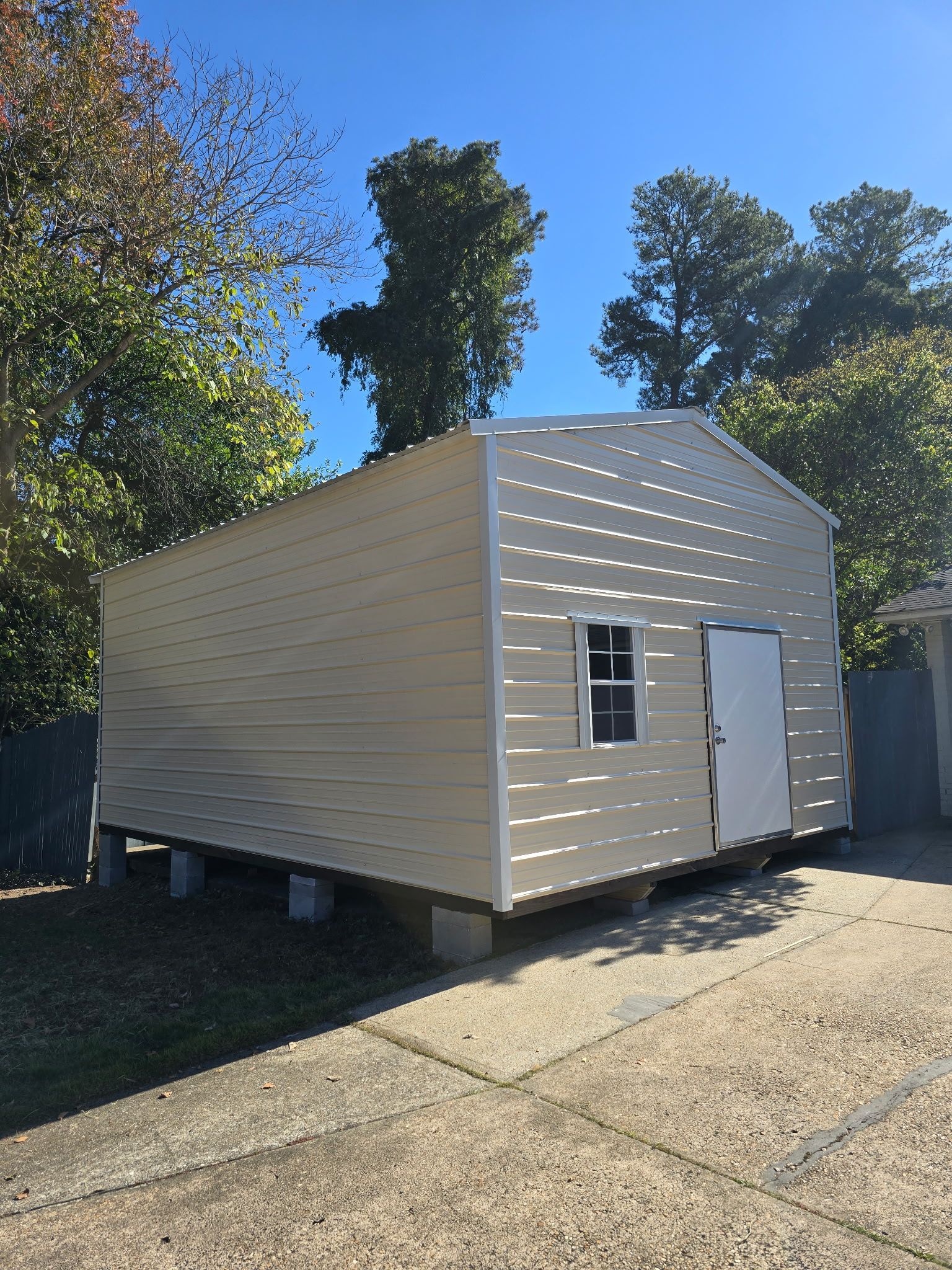 Tan shed on cinder blocks with a white door and small window, set in a yard.