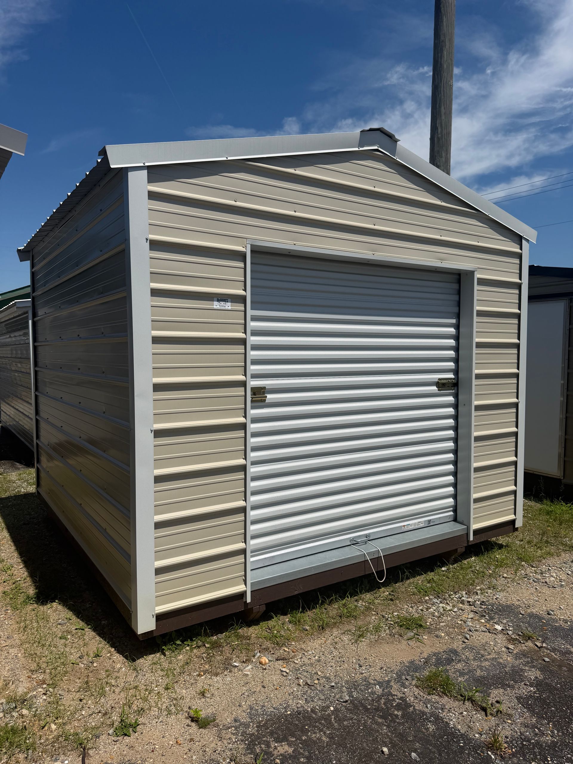 Small beige metal storage shed with a white roll-up door on a dirt lot under a blue sky.