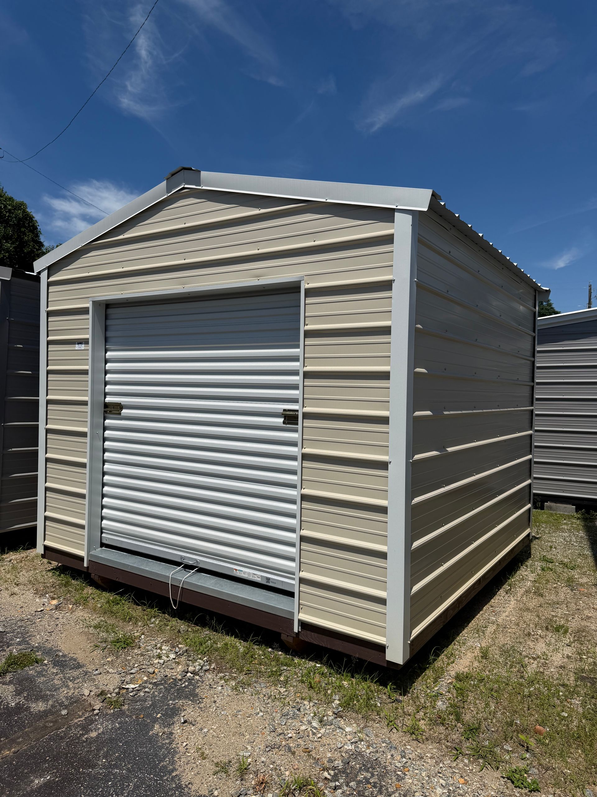 Small beige storage shed with a white roll-up door on a gravel lot under a blue sky.