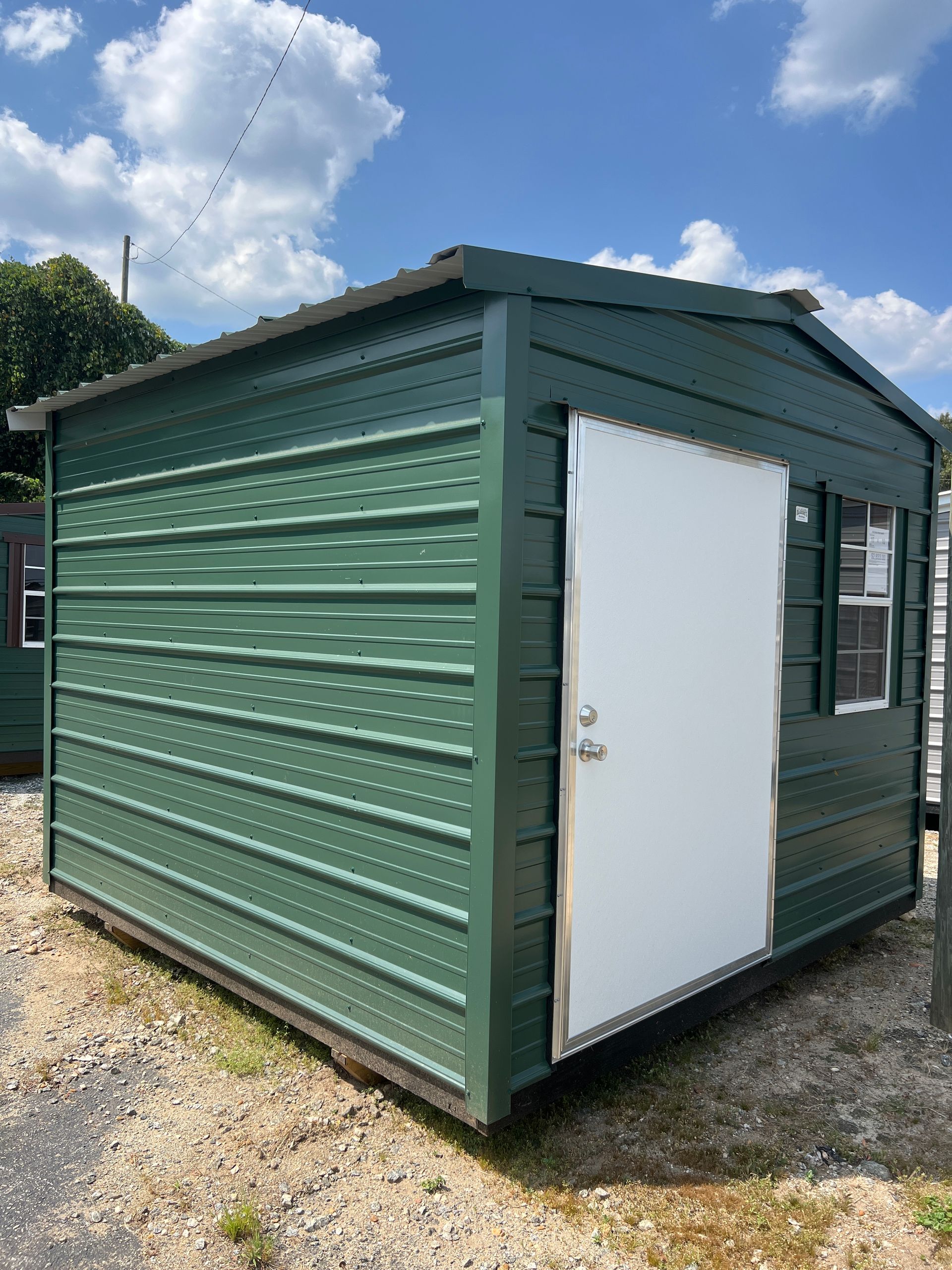 Green metal shed with white door and window, sitting outside on a gravel lot.