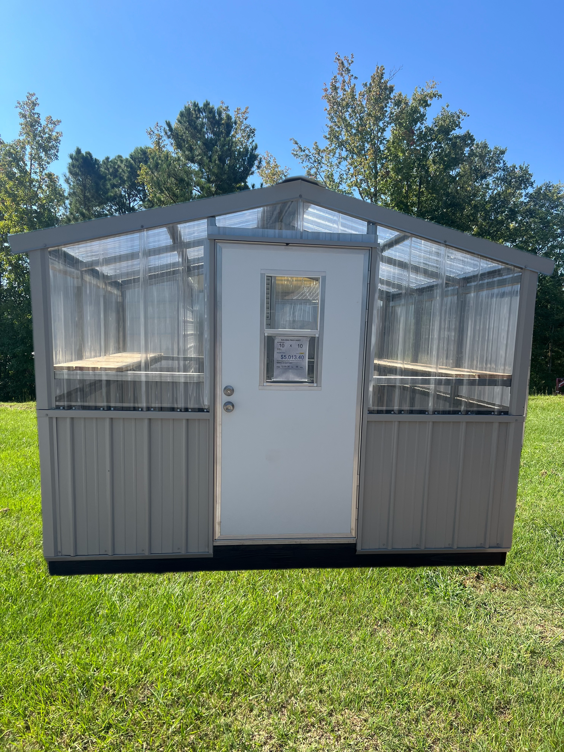 A grey shed-style greenhouse with a white door and translucent, corrugated plastic walls and roof, set on a grassy lawn.