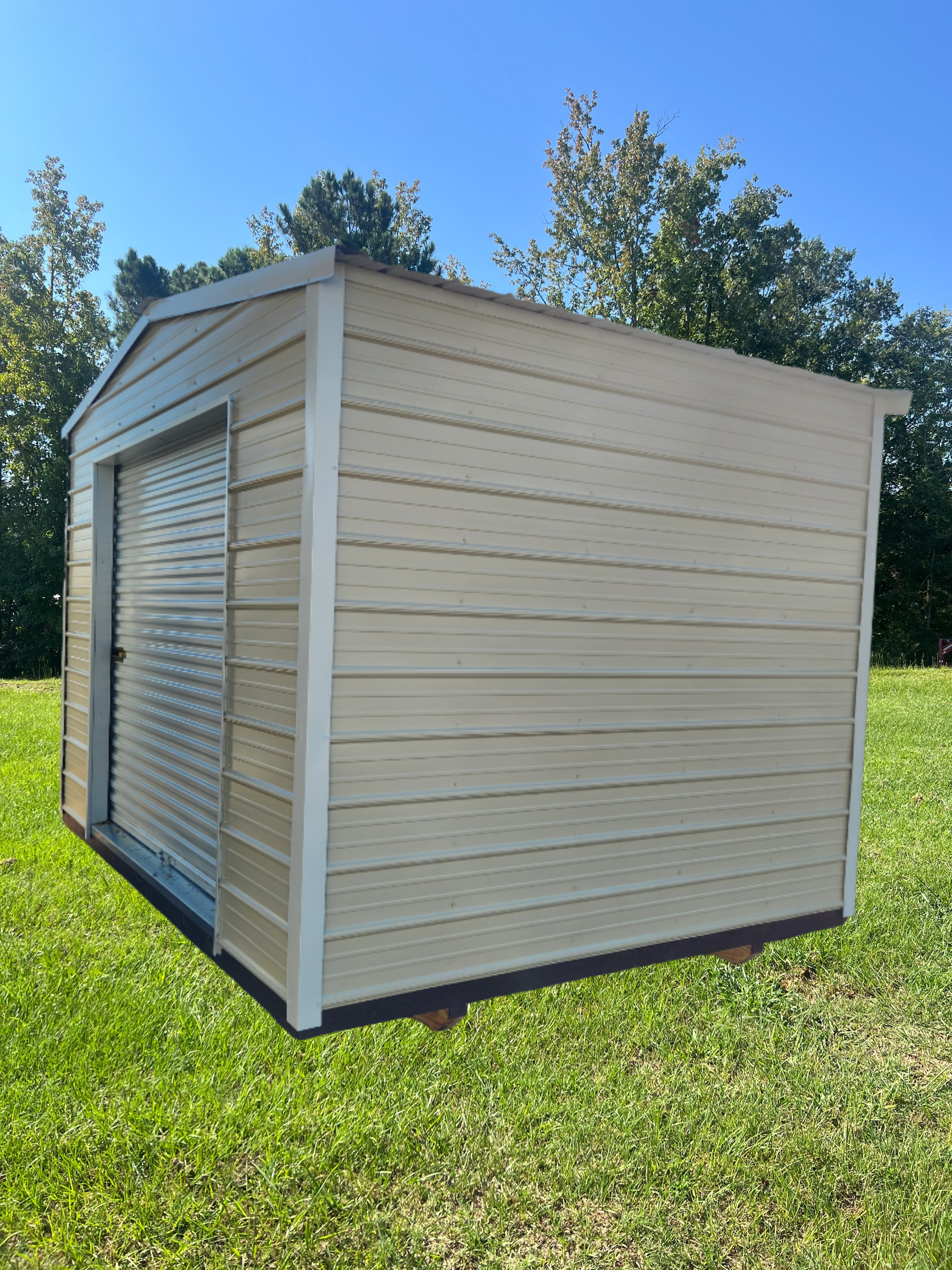 Tan metal shed with roll-up door, on green grass, trees and blue sky background.