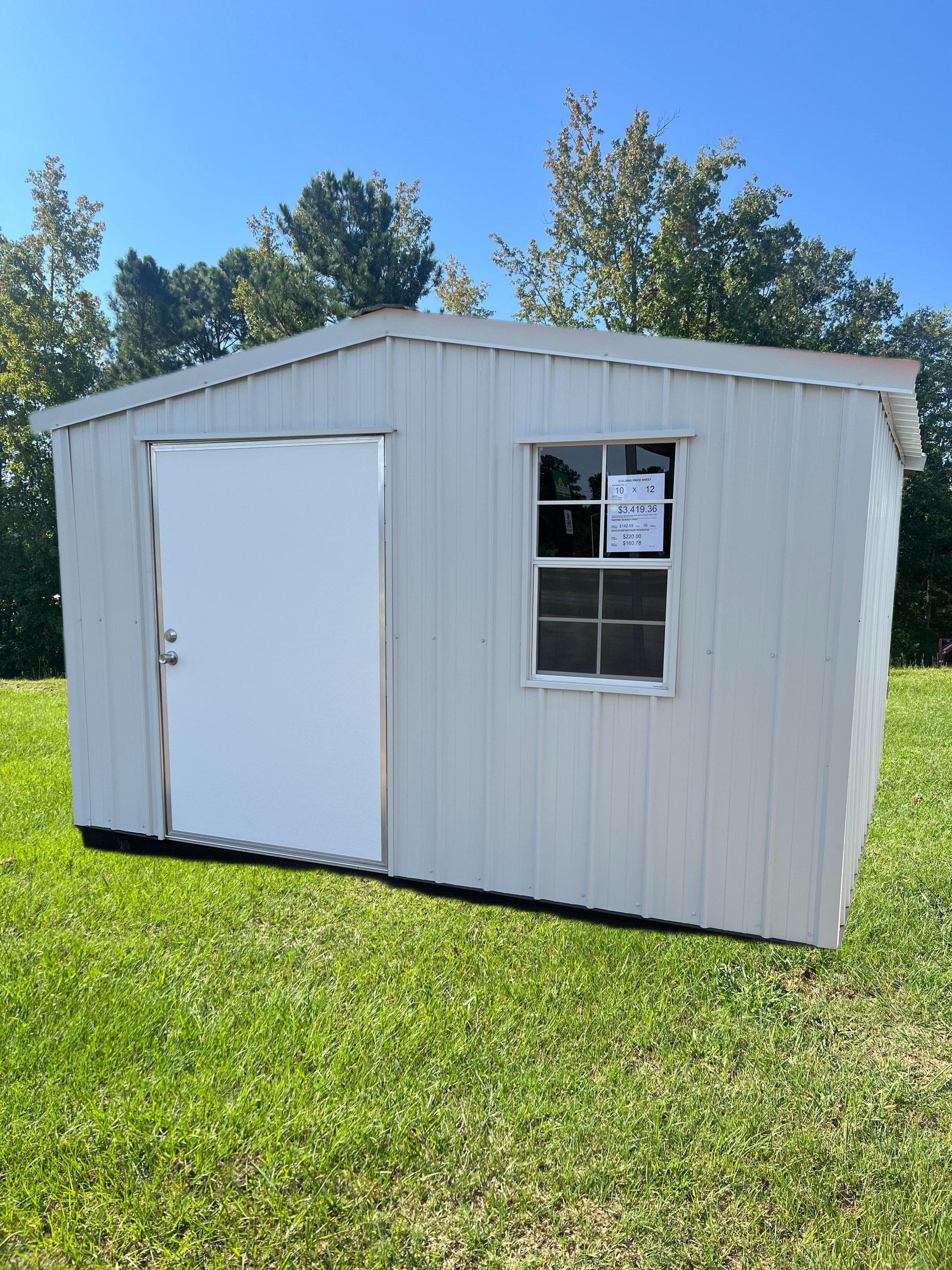 A light gray storage shed with a white door and a small window, sitting on green grass against a clear blue sky.
