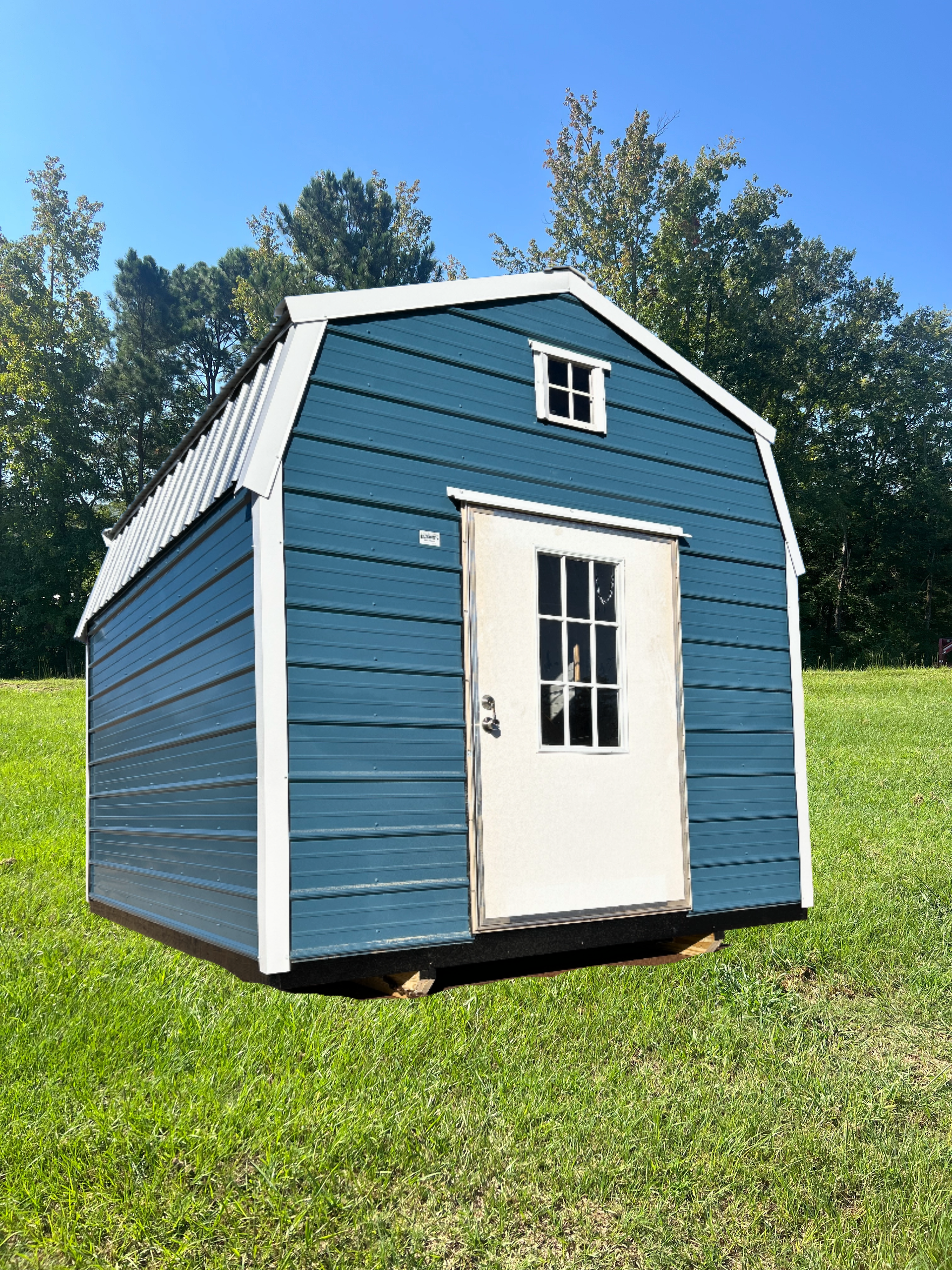 Blue barn-style shed with white trim and door on a grassy field with trees in the background.