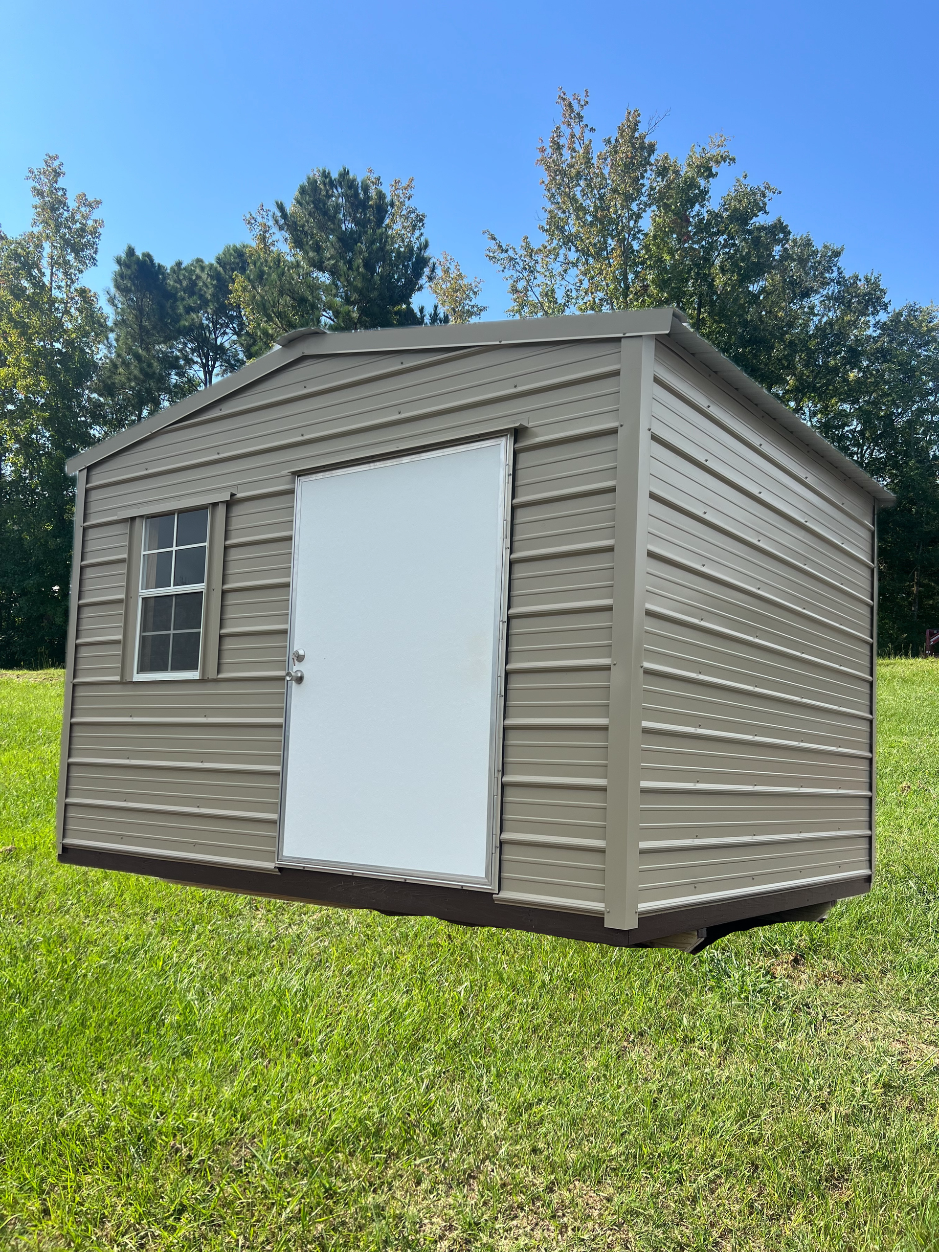 A beige, metal-sided storage shed with a white door and a single window sitting on a grassy lawn under a clear blue sky.