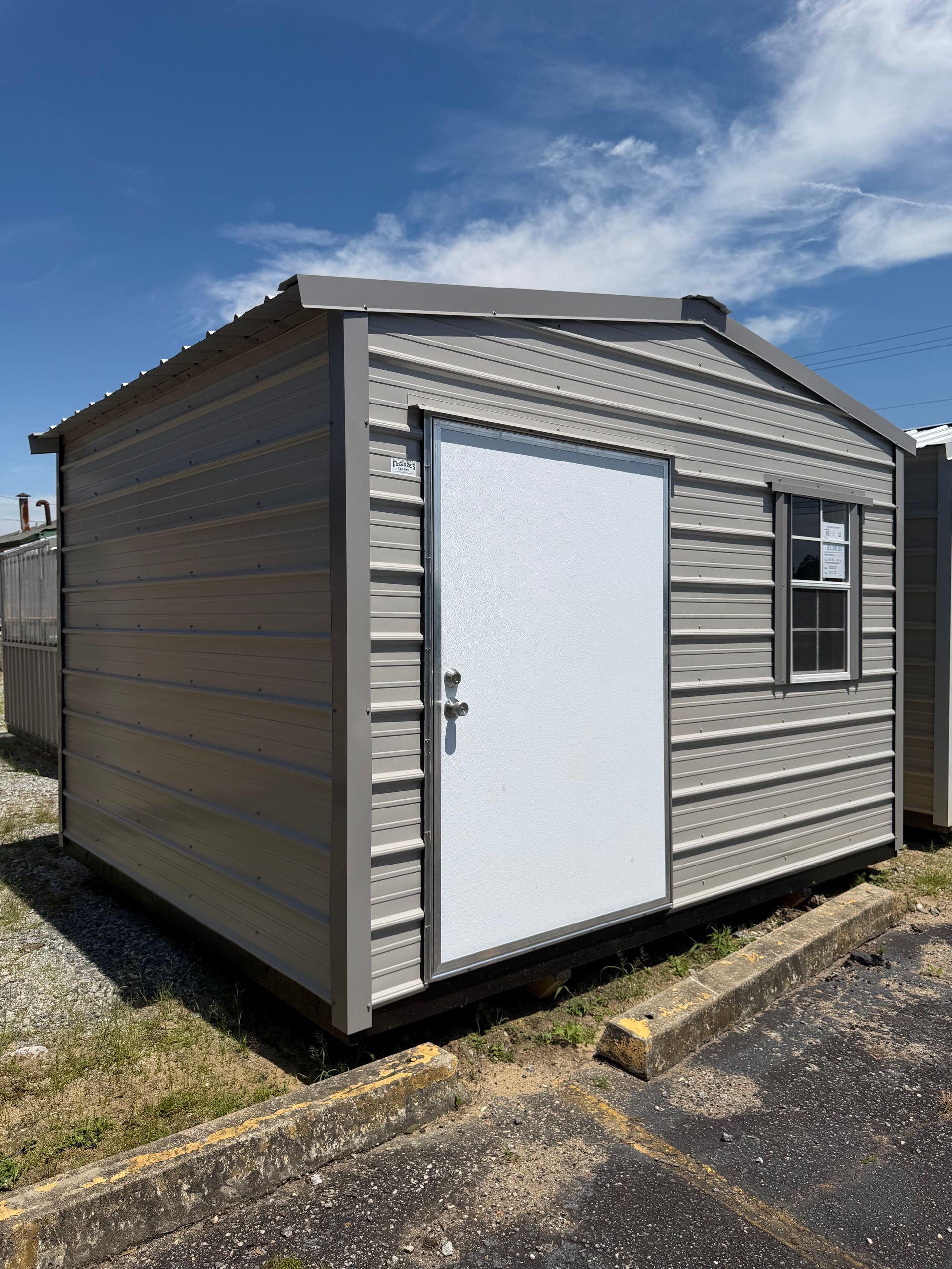 Small beige metal shed with a white door and window on a gravel lot under a blue sky