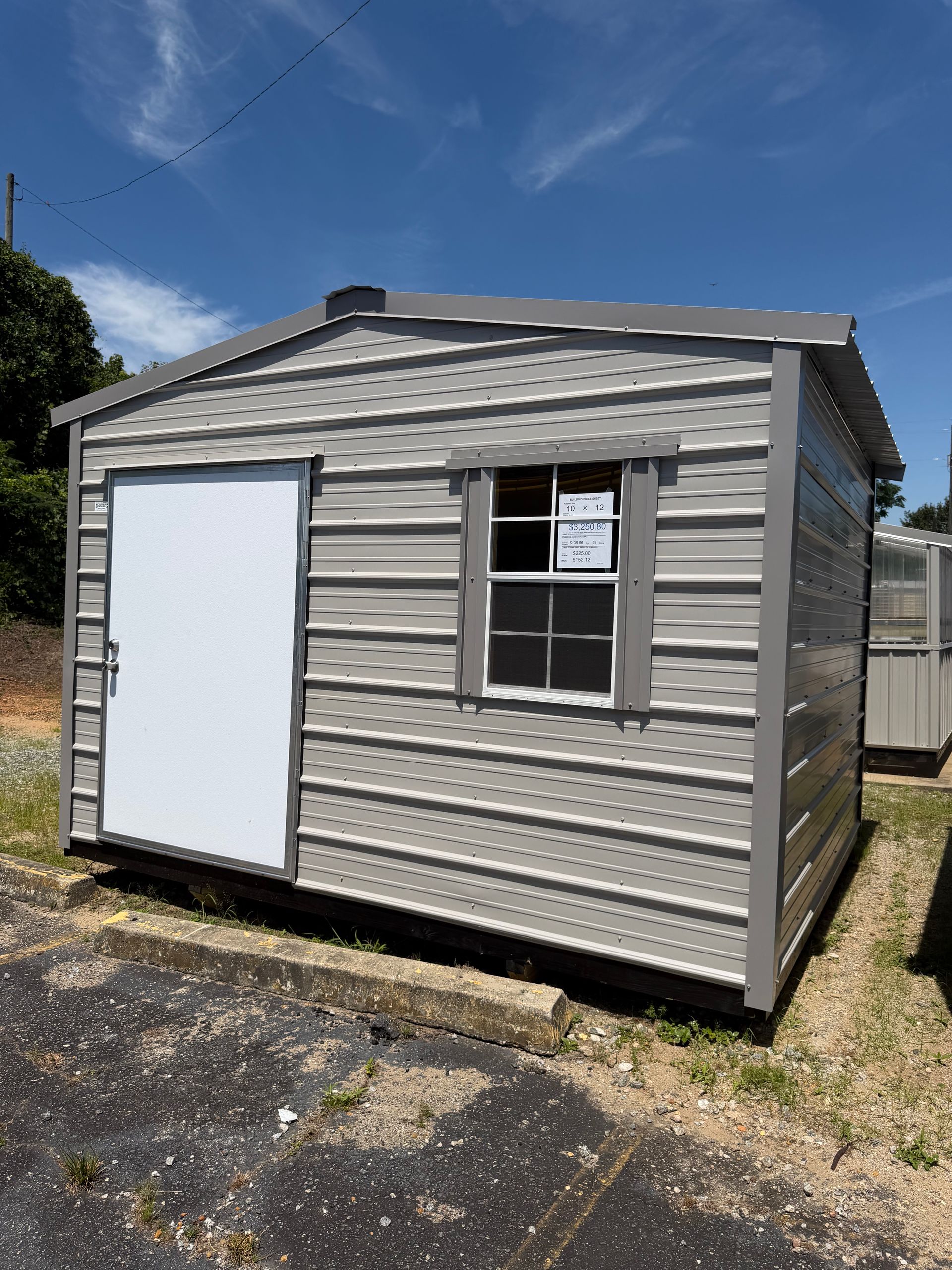 Small gray shed with white trim, a window, and a large white door on a gravel lot under a blue sky