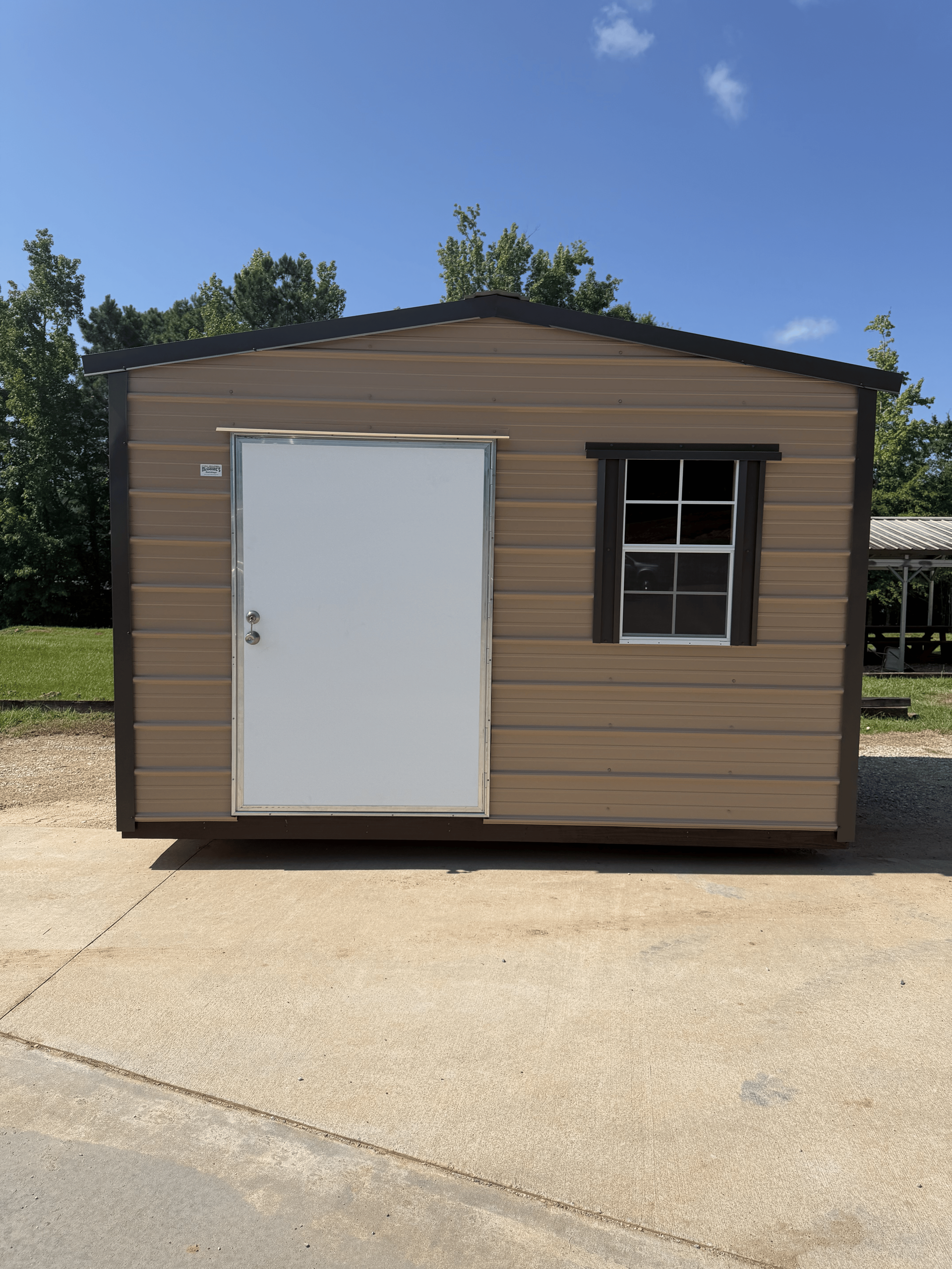 Small tan shed with a white door and window on a concrete driveway under a blue sky