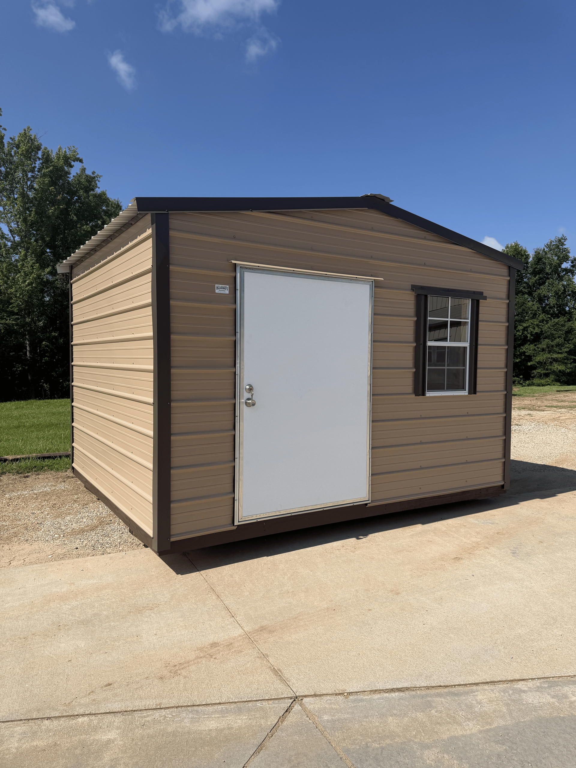 Small tan shed with white door and window sitting on a concrete pad under a blue sky.