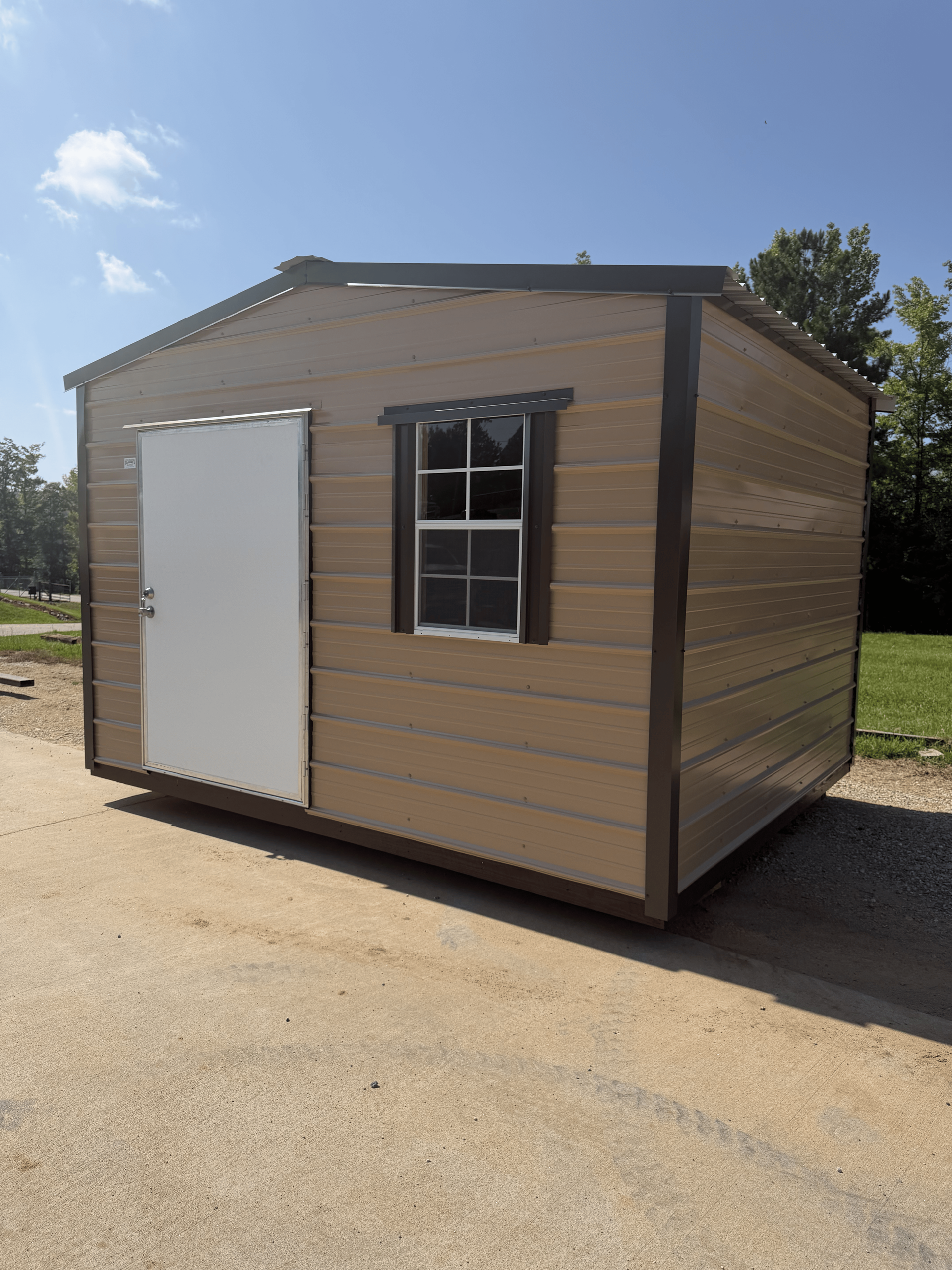 Small tan shed with a white door and window, standing on a paved lot under a blue sky.