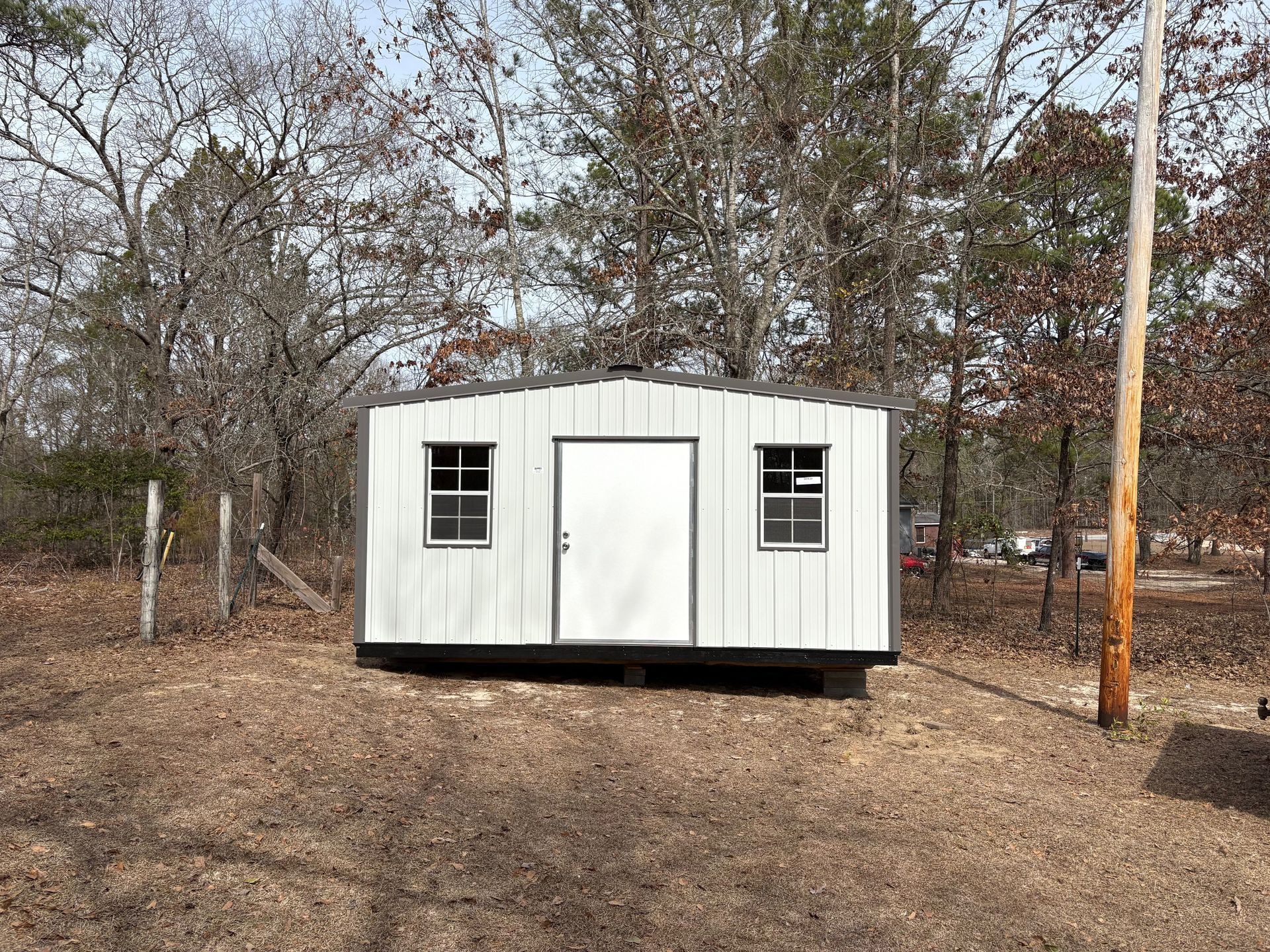 White shed with two windows and a door, set in a wooded area.
