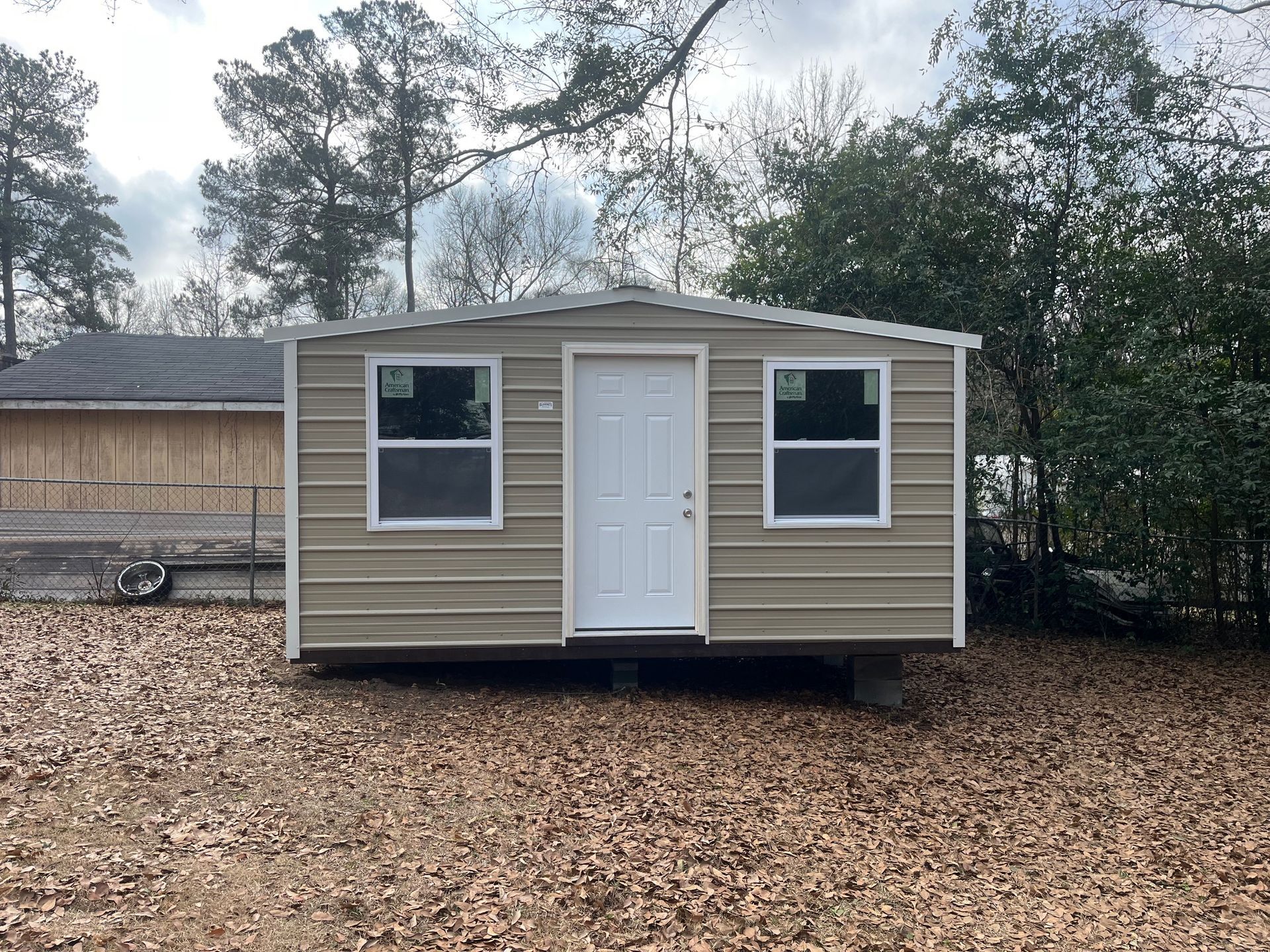 Tan metal storage building with white door and windows; set on a brown leaf-covered ground.