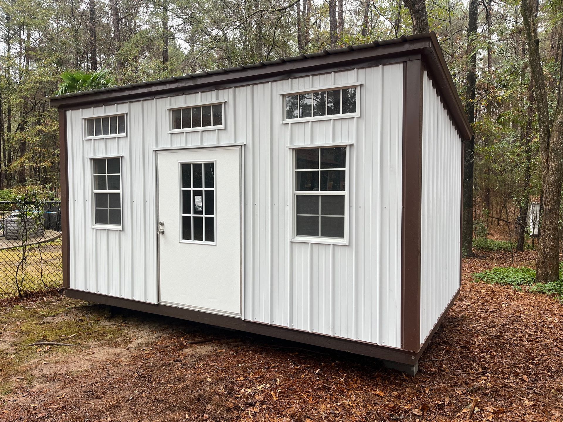 White and brown shed with windows and a door in a wooded area.