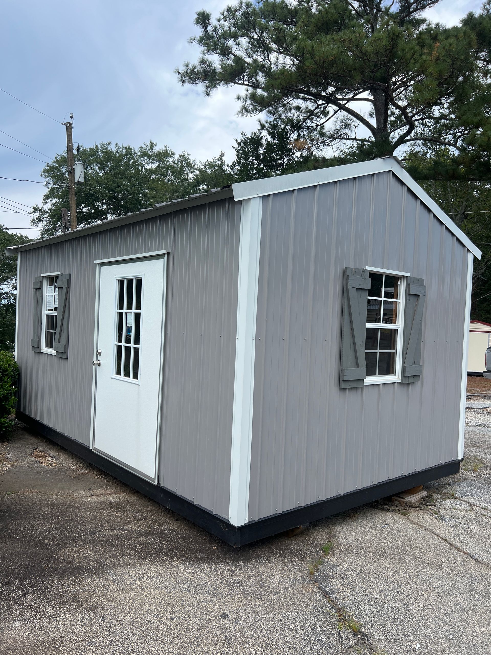 Grey shed with white trim, a white door, and two windows with gray shutters. Outdoors.