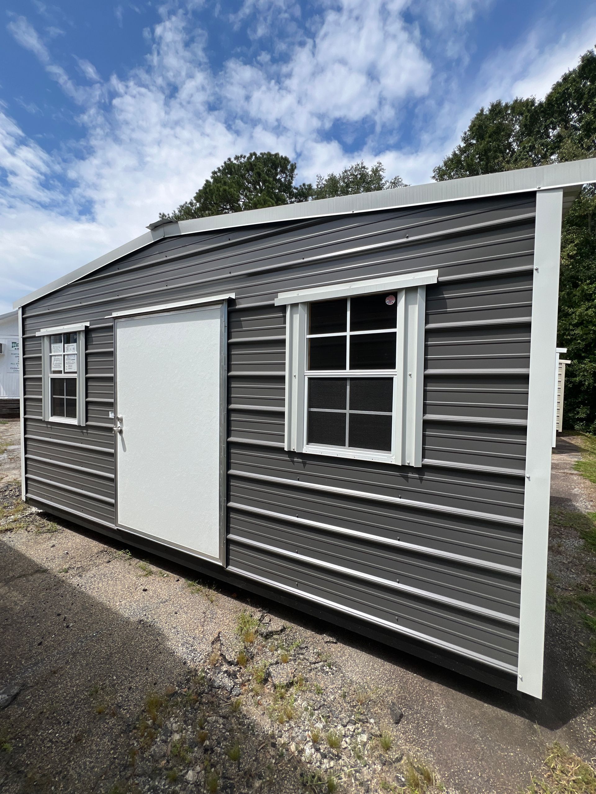 Gray metal shed with a white door and two windows, under a partly cloudy sky.
