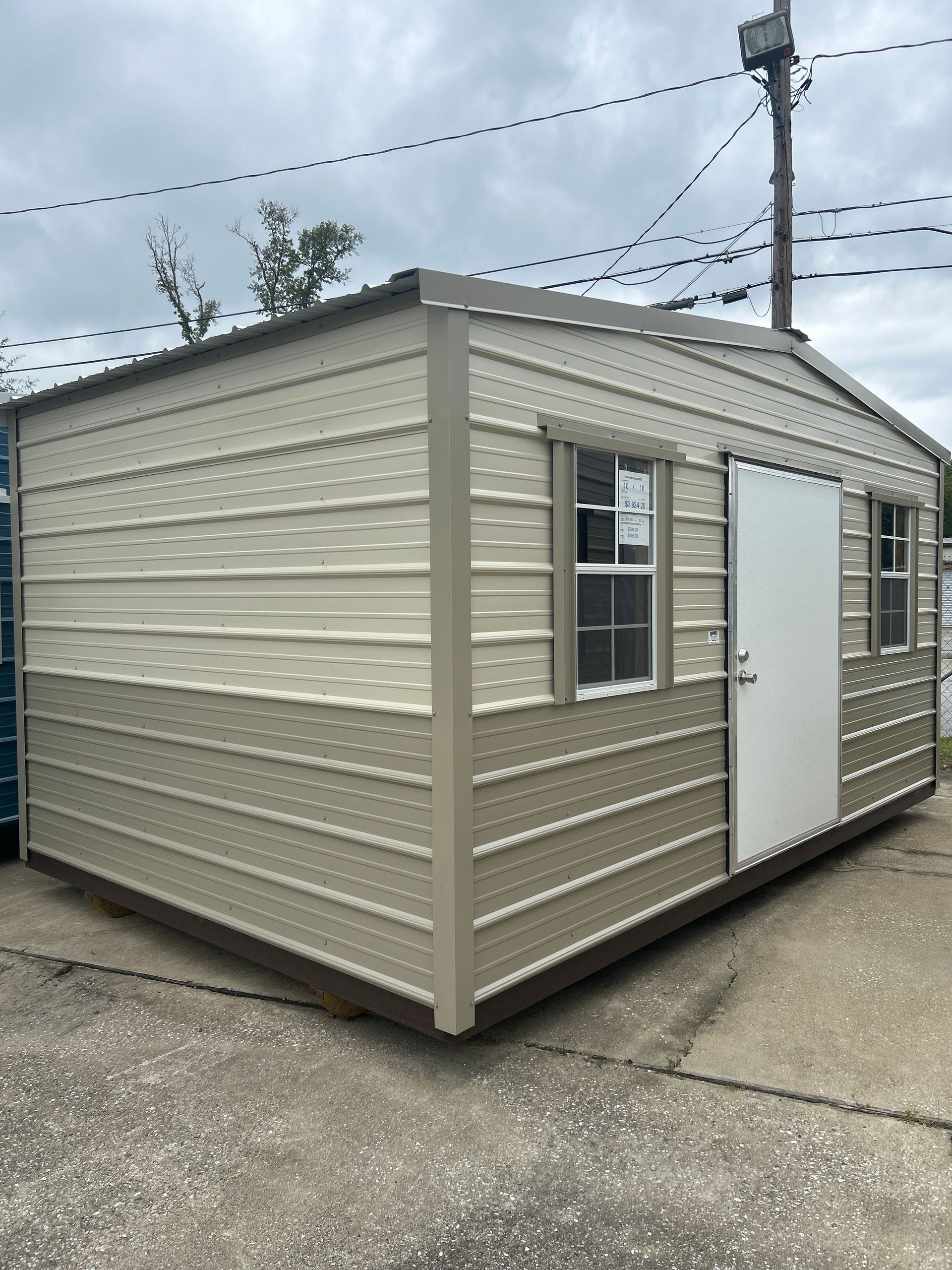 Tan metal storage shed with a white door and two windows.