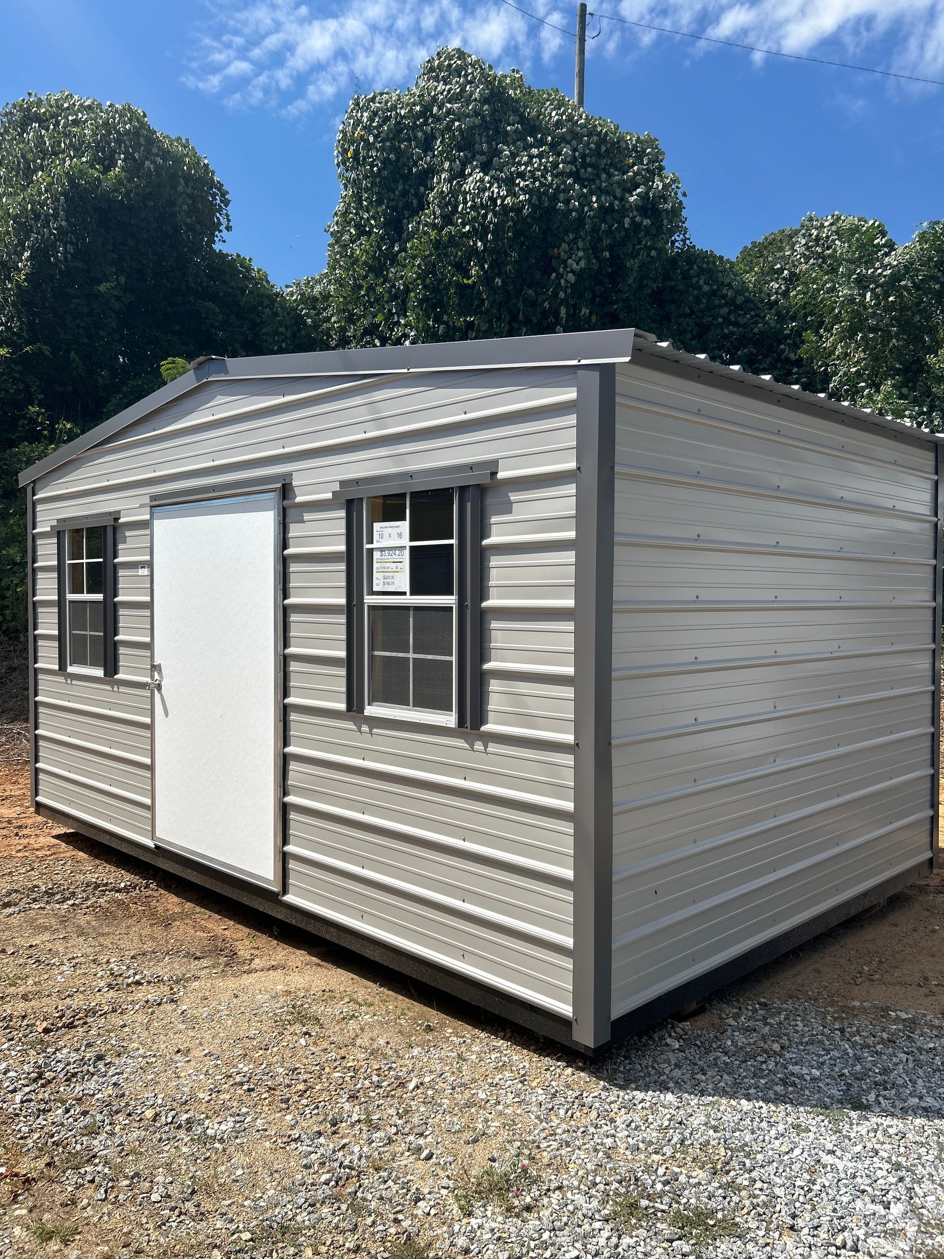 Gray metal shed with white door and windows, outdoors on a gravel surface.