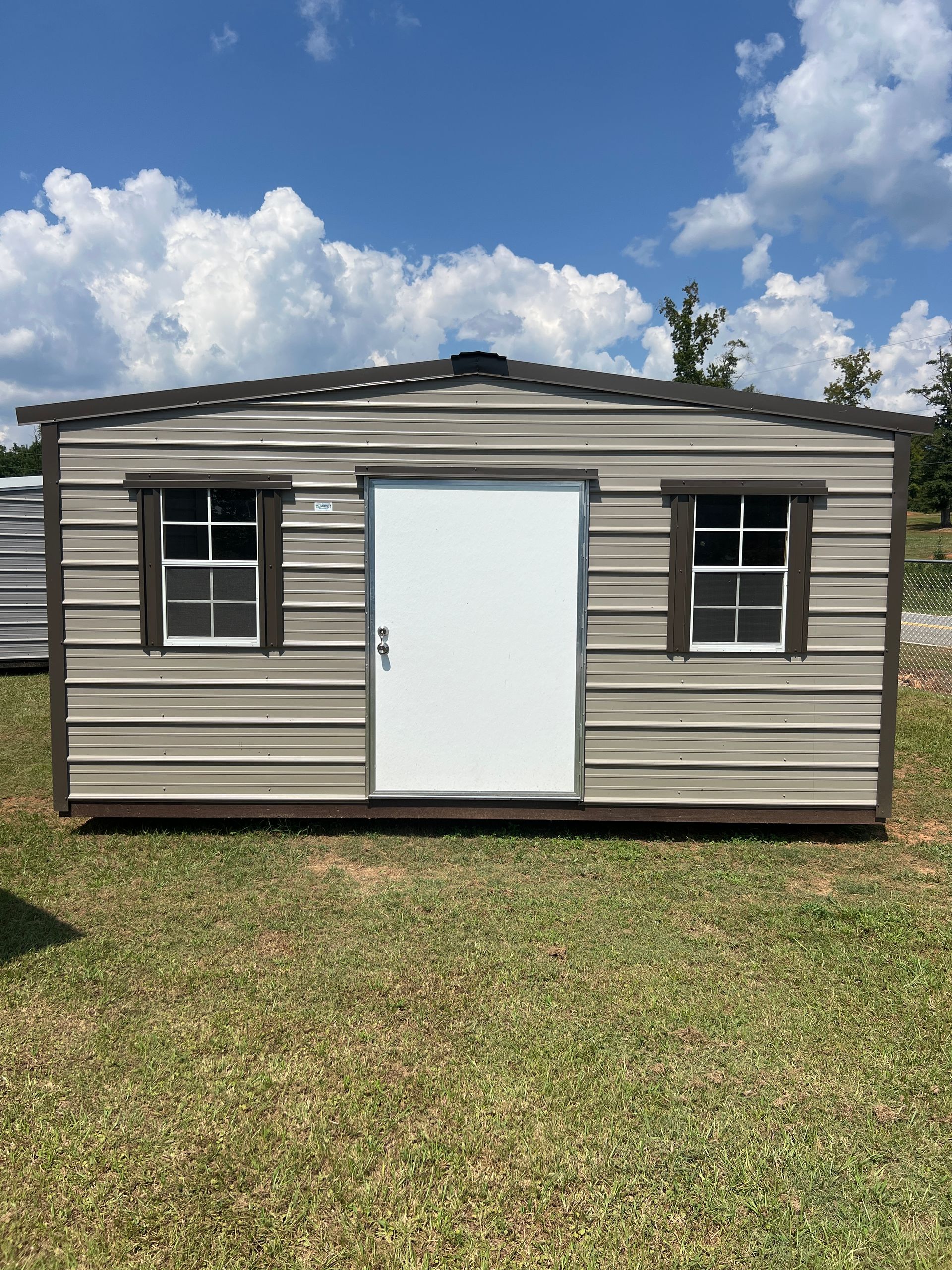 Tan metal storage shed with a white door and two windows, set outdoors on grass under a blue sky.
