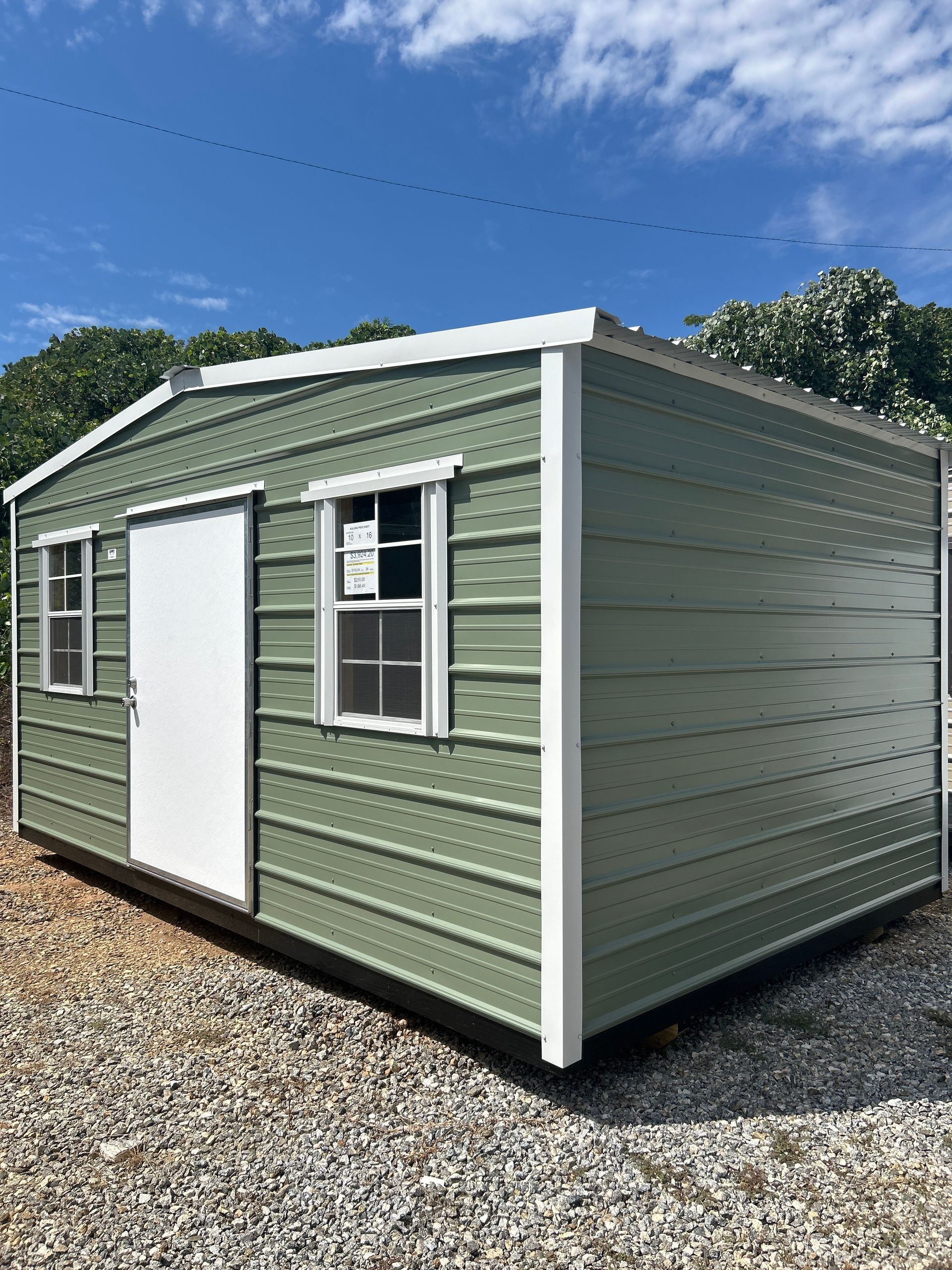Green metal storage shed with white trim, door, and windows. Outdoors, gravel ground, blue sky.