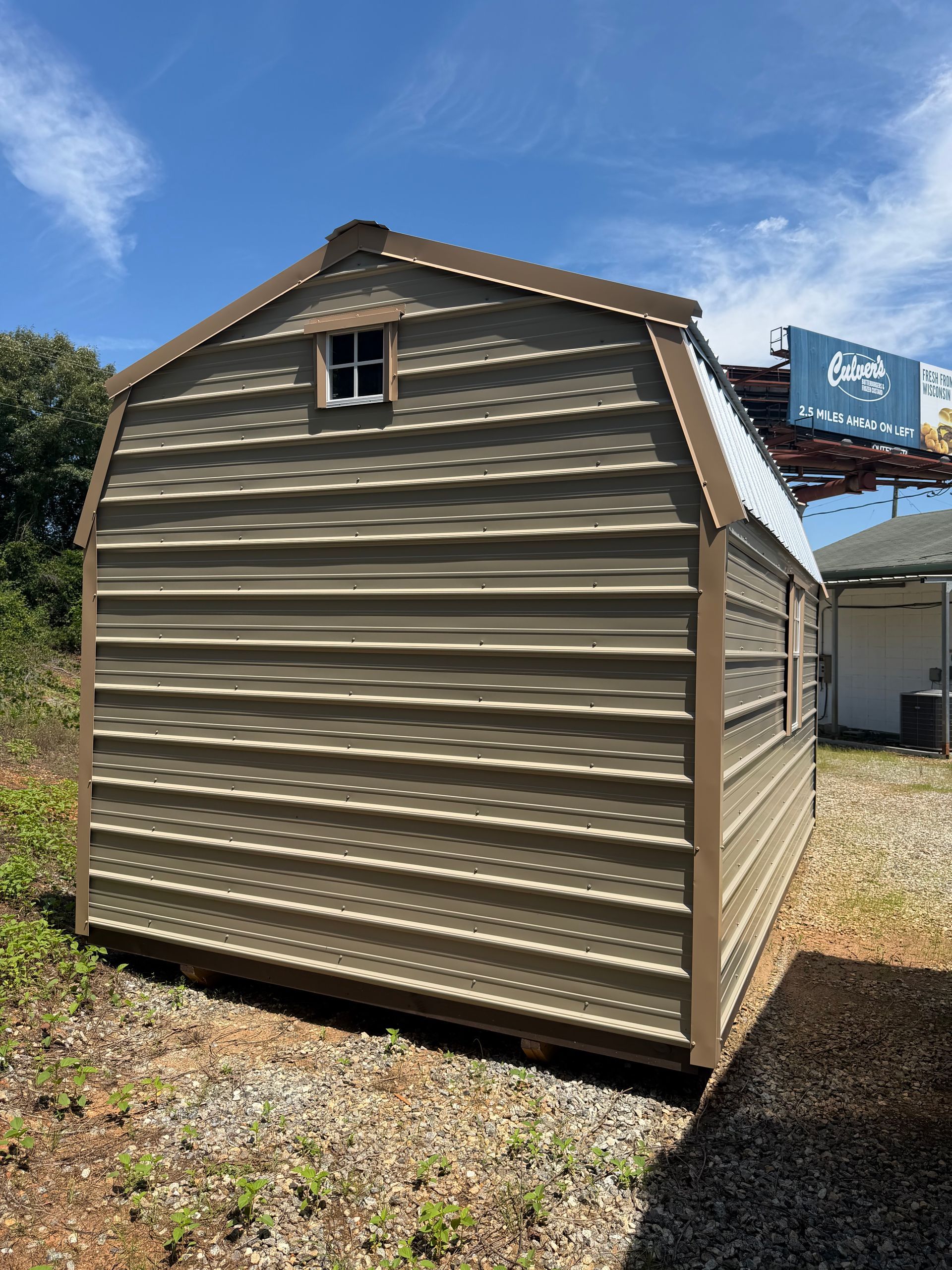 Beige metal storage shed with a small window, sitting on gravel under a blue sky.