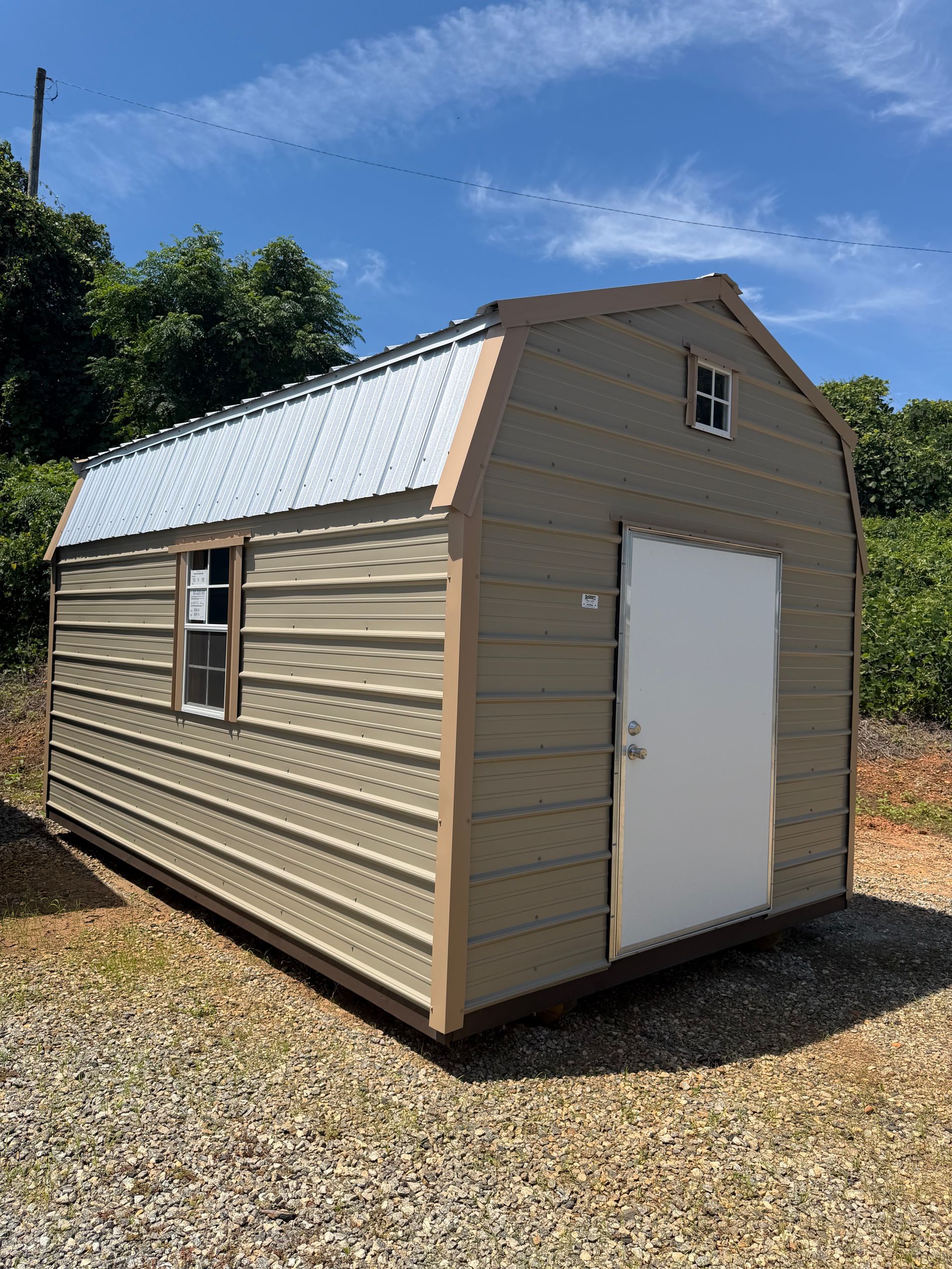 Small tan shed with a white door and metal roof, sitting on gravel under a blue sky.