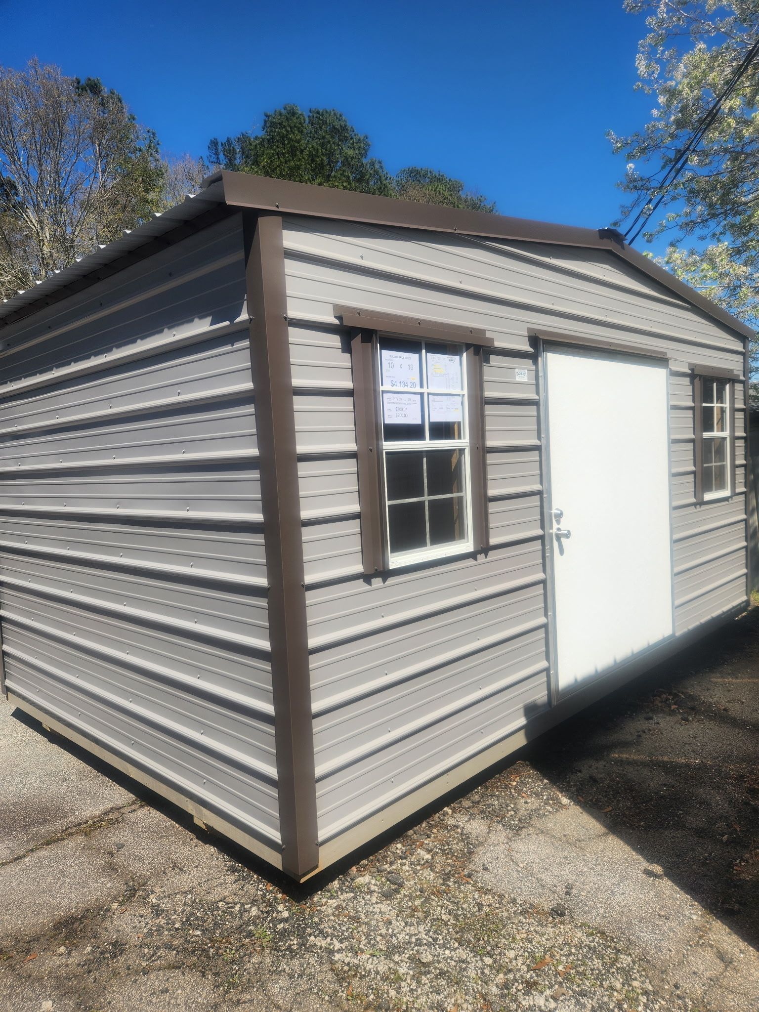 A light grey metal storage shed with brown trim, a white door, and two windows, situated on a gravel surface outdoors.