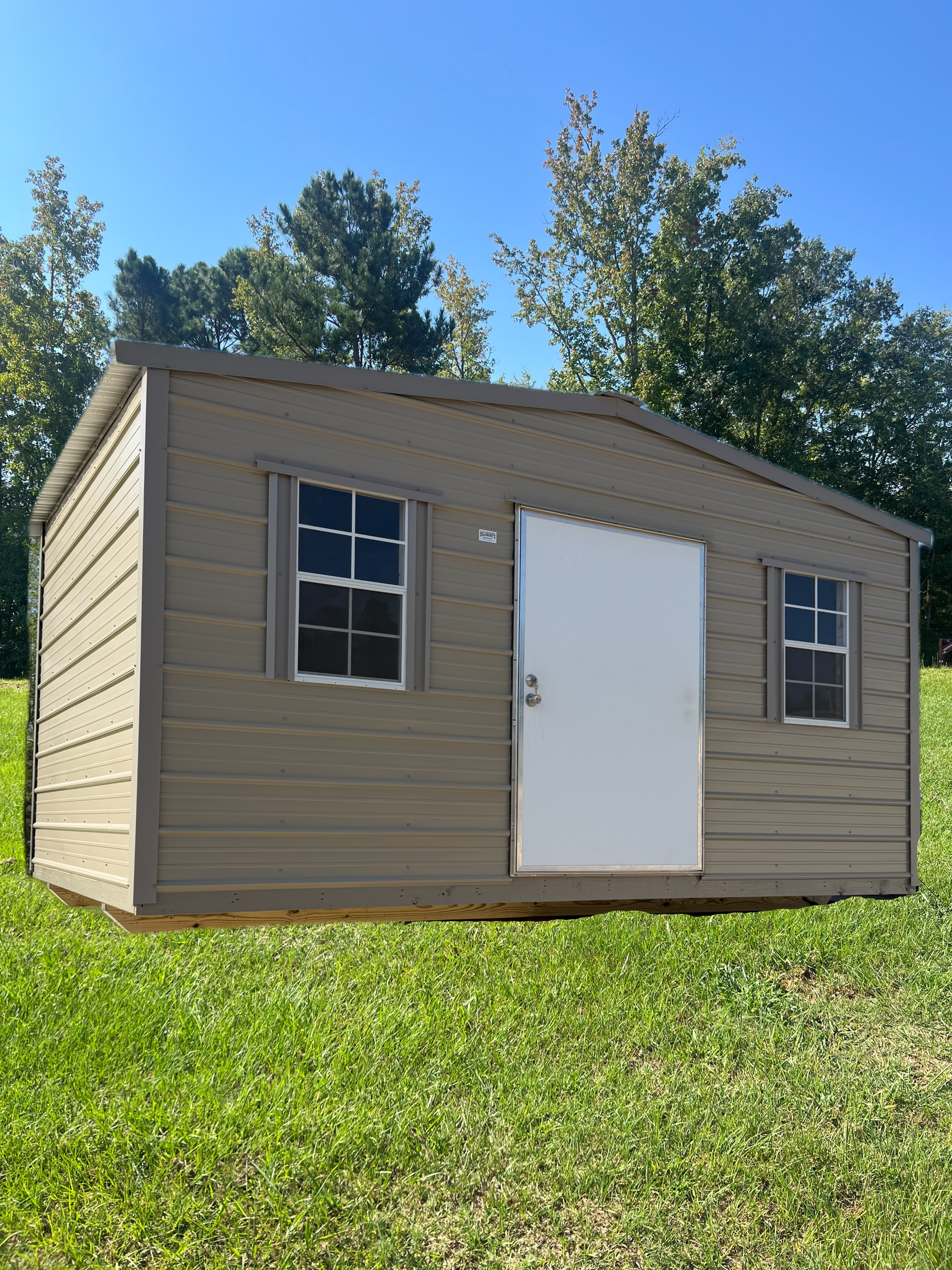 A beige storage shed with a central white door and two side windows sits on a grassy lawn under a clear blue sky.