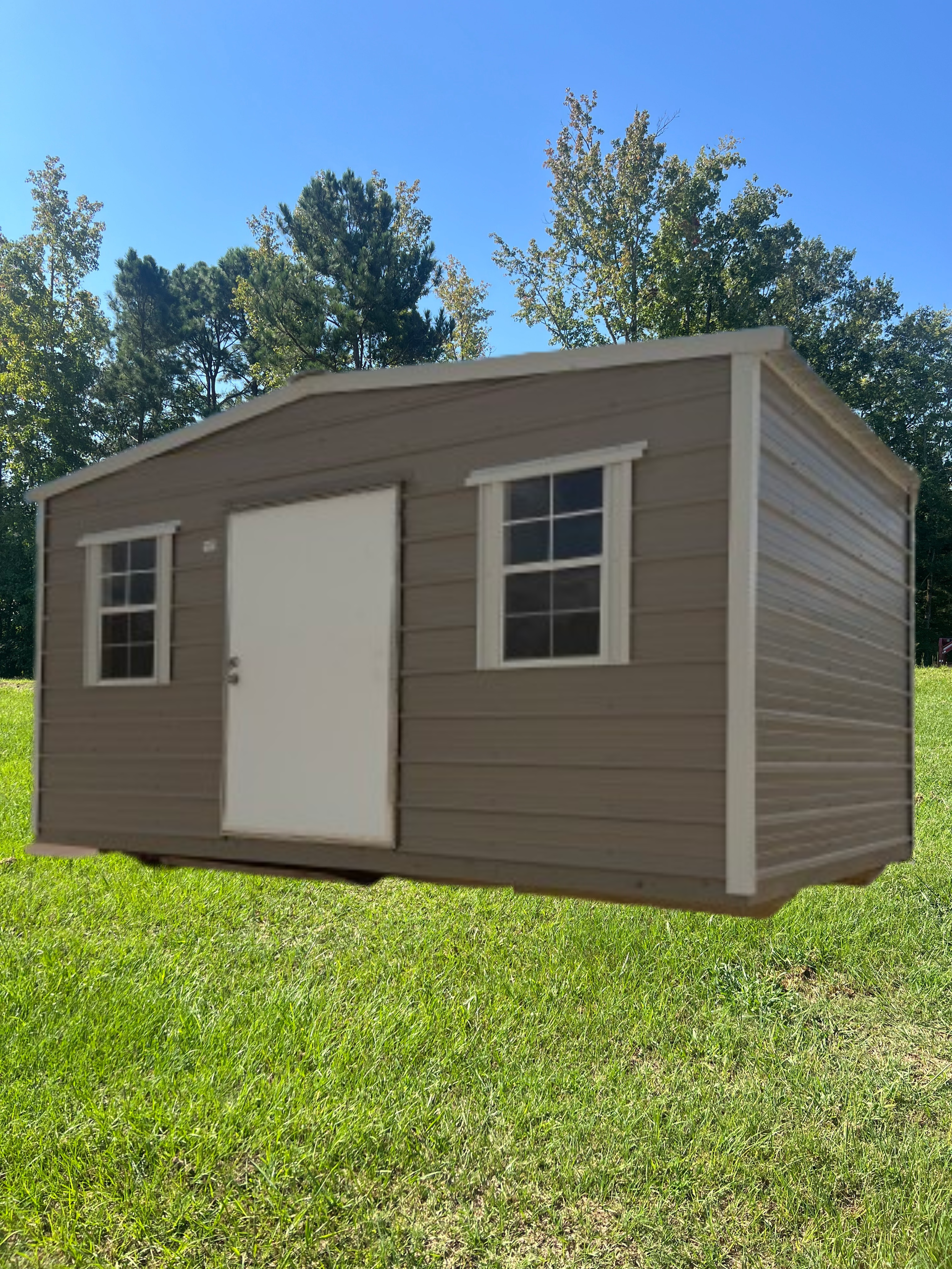 A tan, rectangular storage shed with a single white door and two windows sitting on a grassy lawn under a blue sky.
