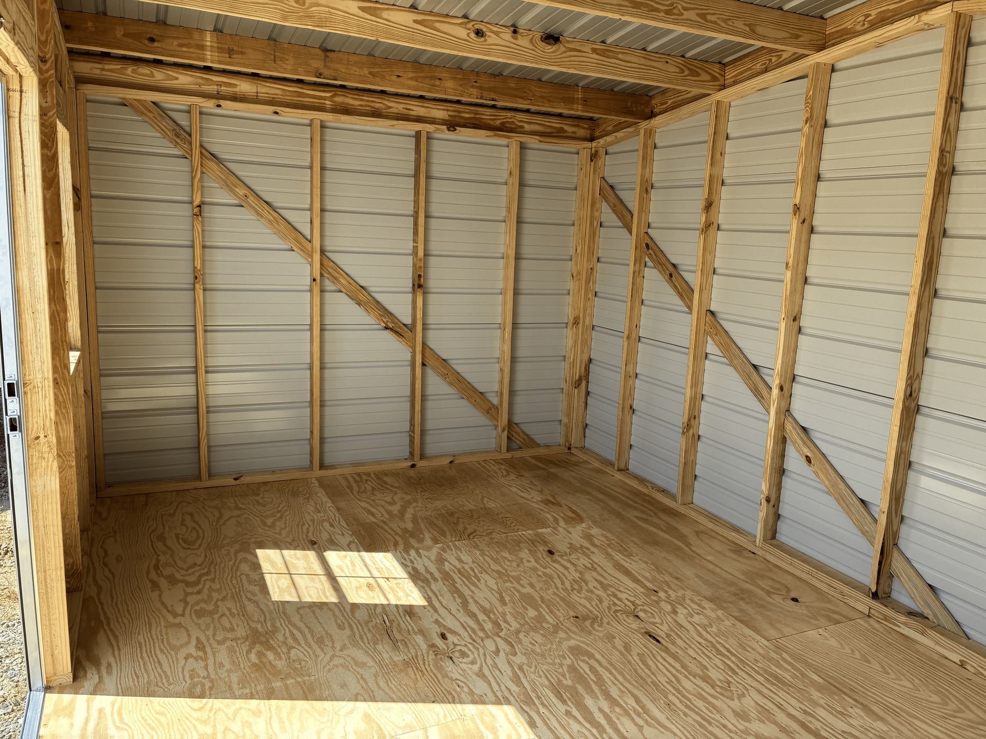 Empty unfinished shed interior with exposed wooden framing and plywood floor, lit by sunlight through a doorway.