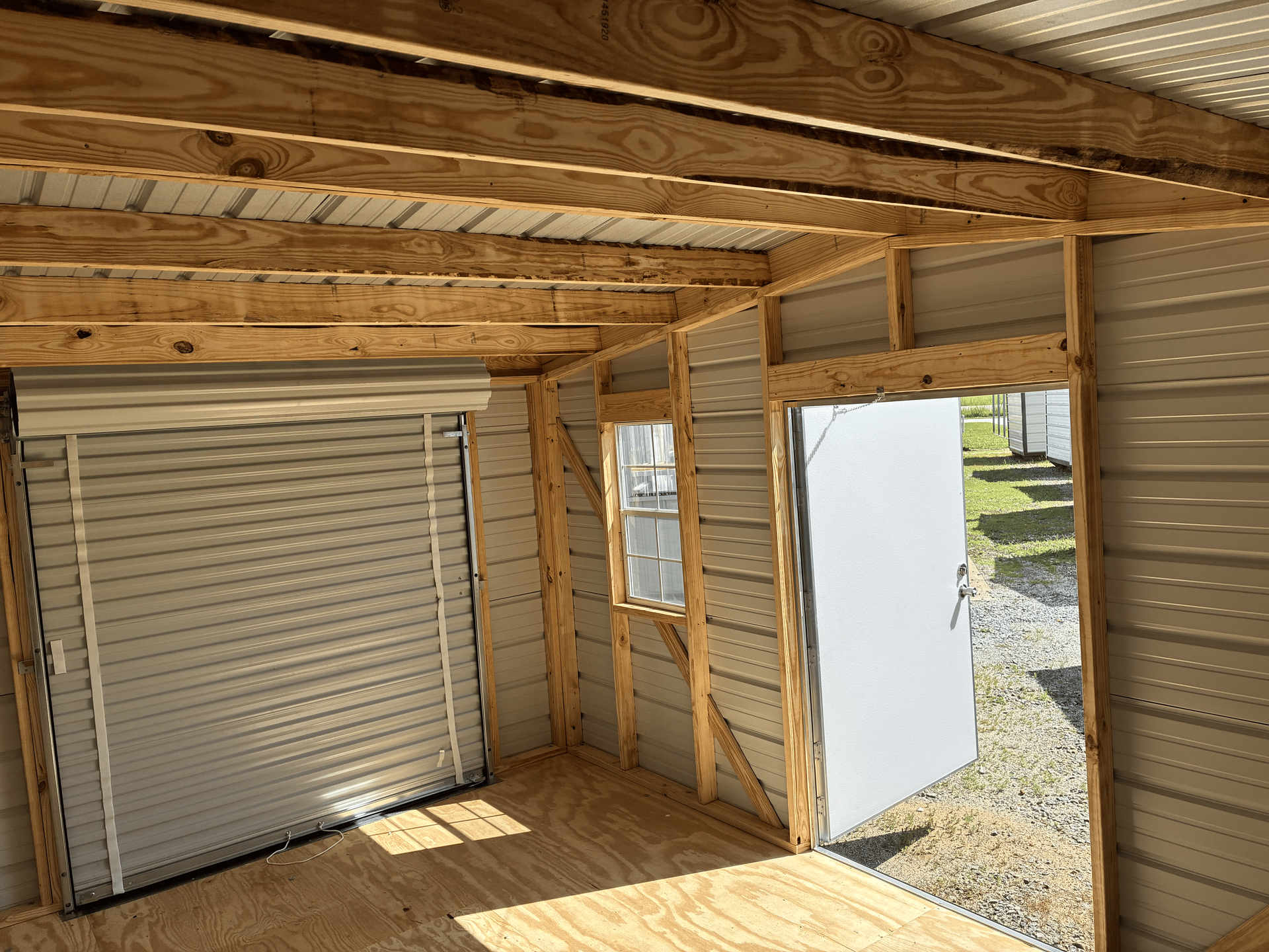 Wood-framed shed interior with roll-up doors, plywood walls, and sunlight on the floor