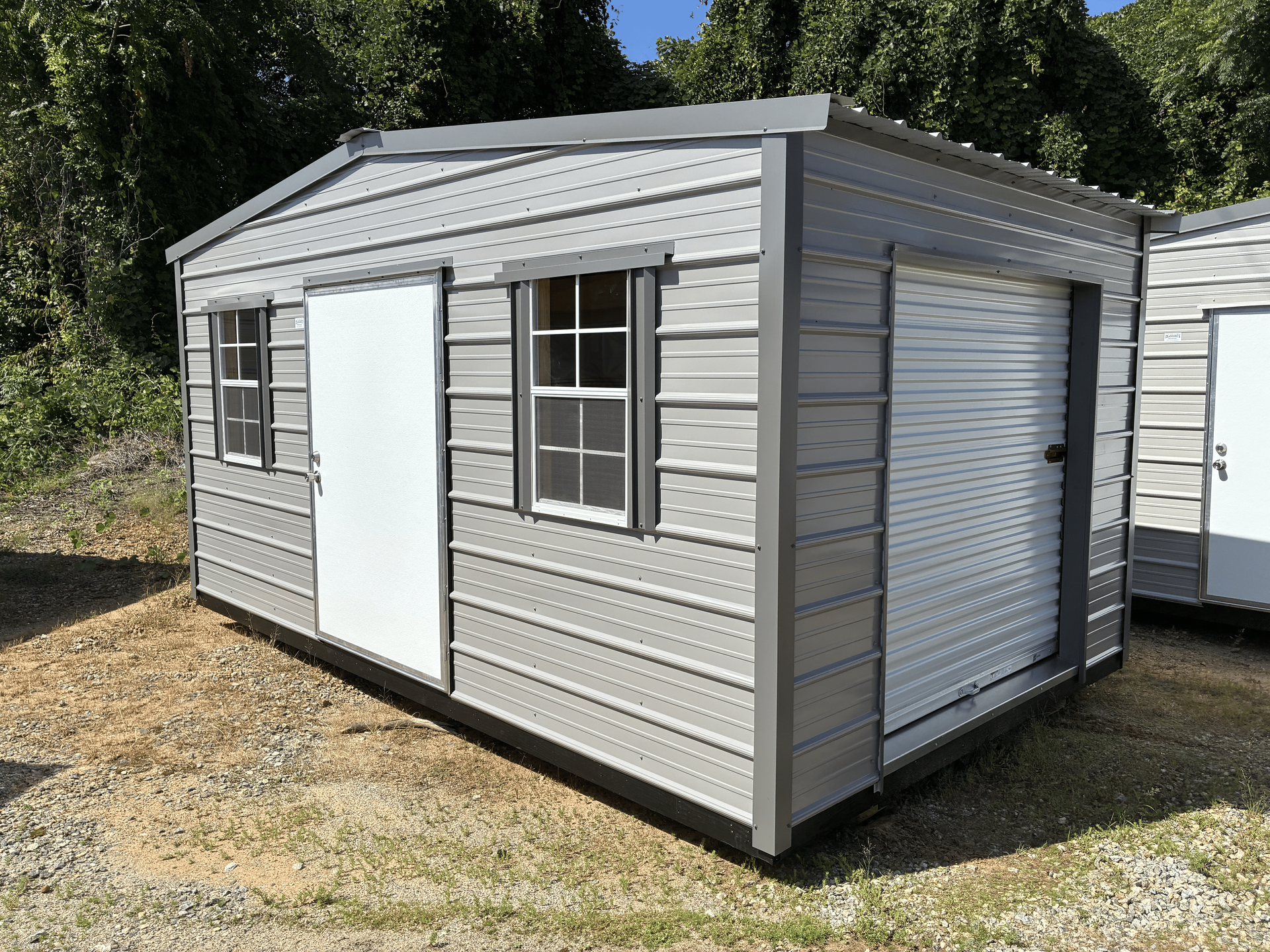 Small gray portable shed with white door and shuttered window on gravel lot