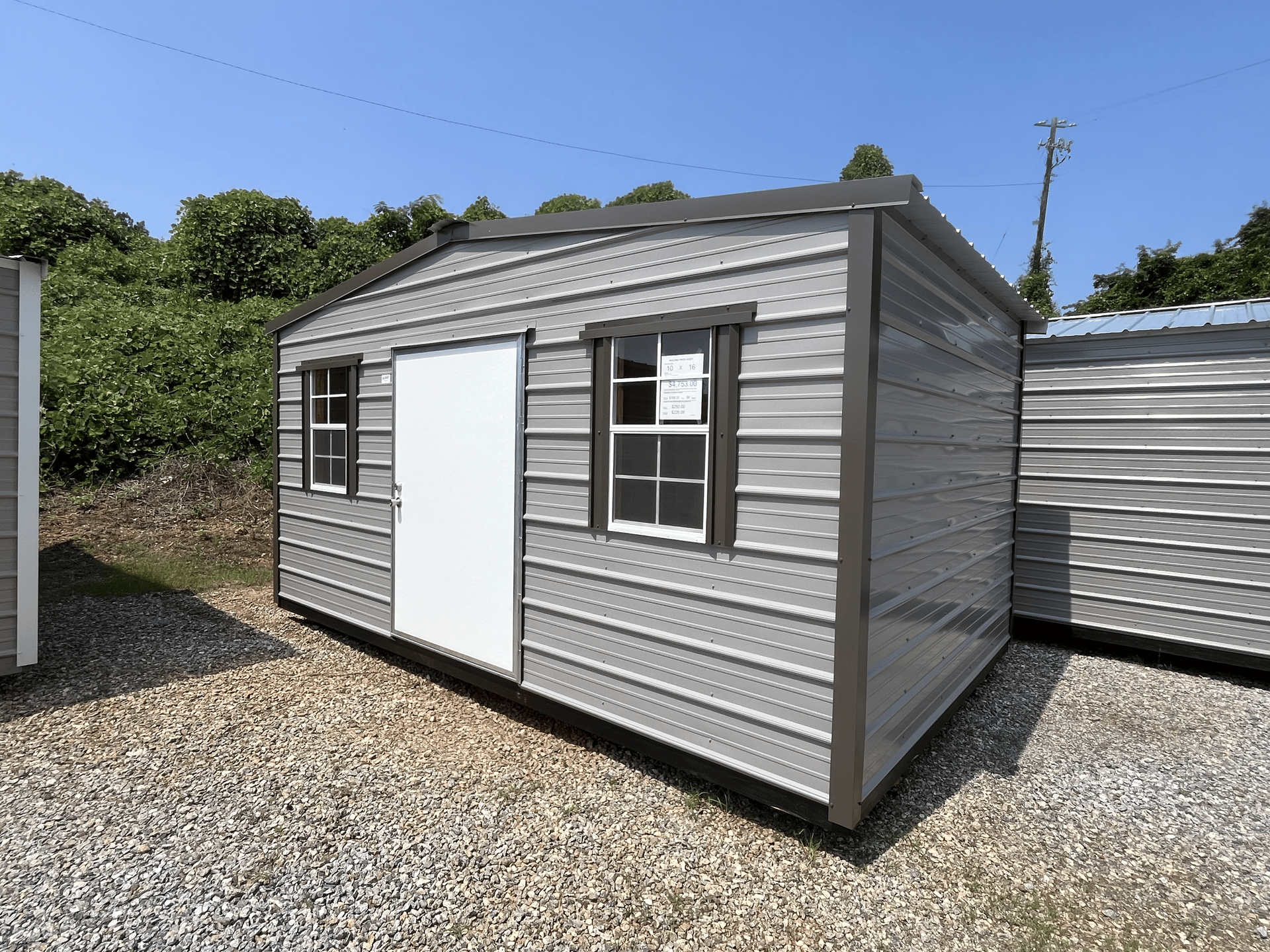 Gray utility shed with white door and window on a gravel lot under a clear blue sky